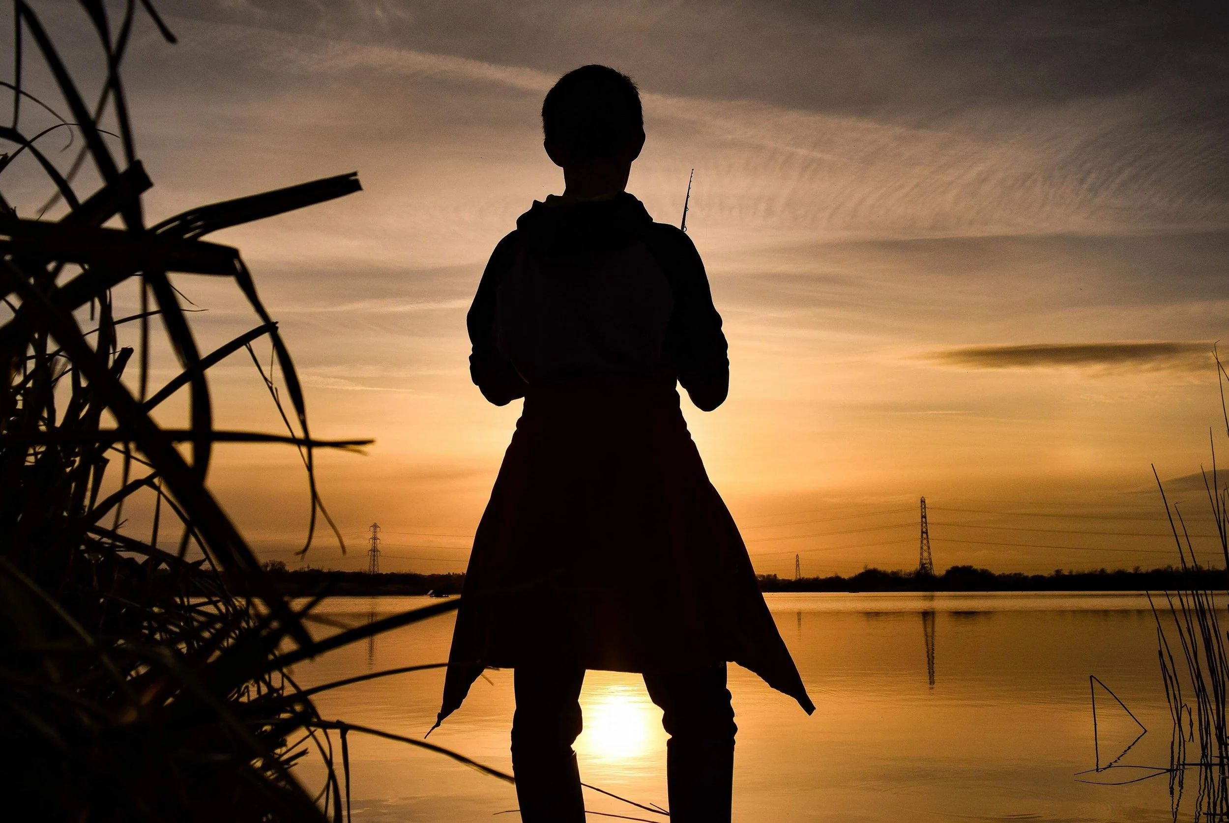 Silhouette of a boy standing near a body of water during sunset, holding a fishing rod, with power lines in the distance and tall grass in the foreground.