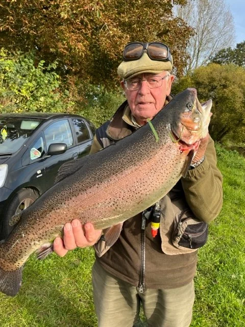 An elderly man in outdoor gear holding a large rainbow trout fish he caught, with a car and trees in the background.