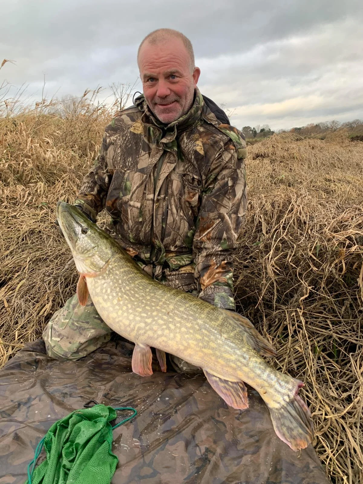 Man dressed in camouflage clothing holding a large fish, sitting on a wet cloth in a marsh, with dried grass and cloudy sky in the background.