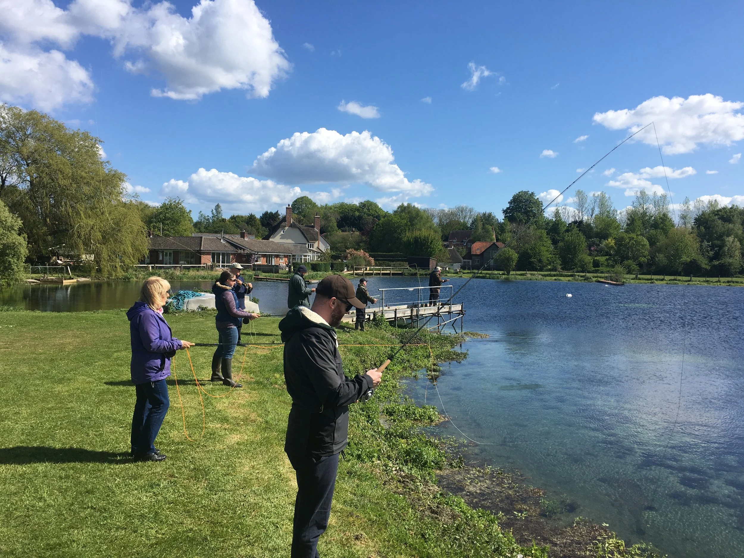 People fishing by a river under a blue sky with scattered clouds and green trees.