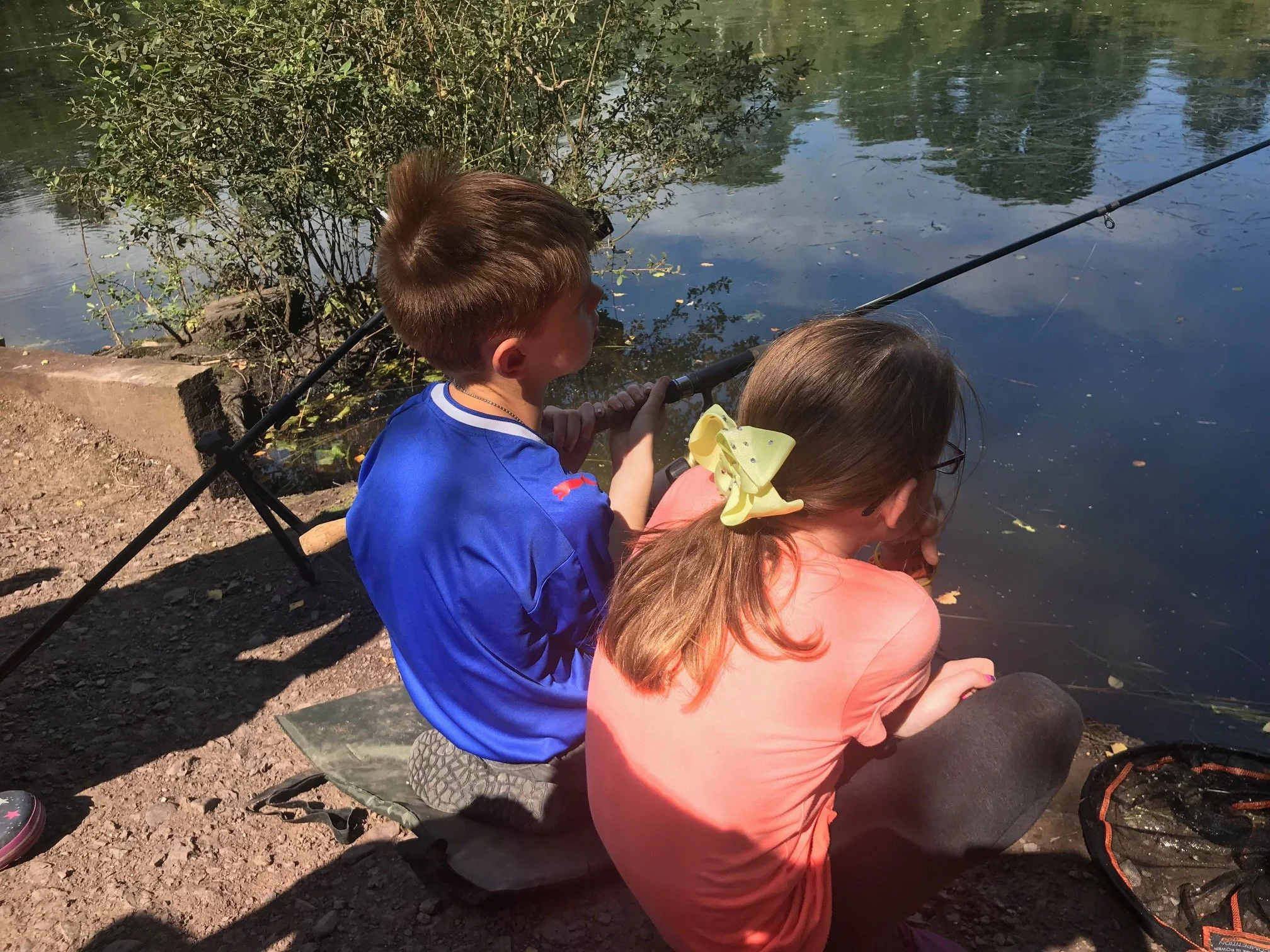 Two children fishing by a pond, sitting on a mat with fishing rods, surrounded by trees and water.