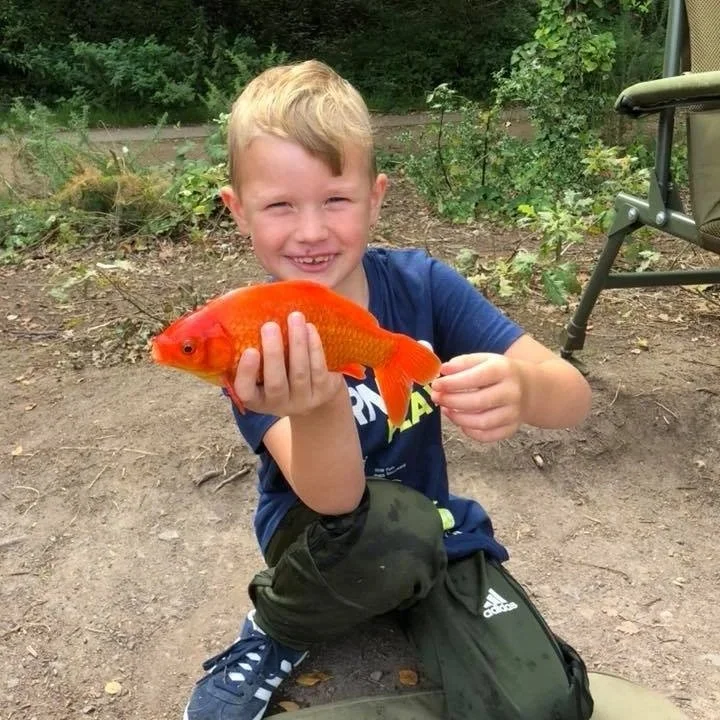 A young boy sitting outdoors on dirt ground, holding a bright orange fish and smiling at the camera.