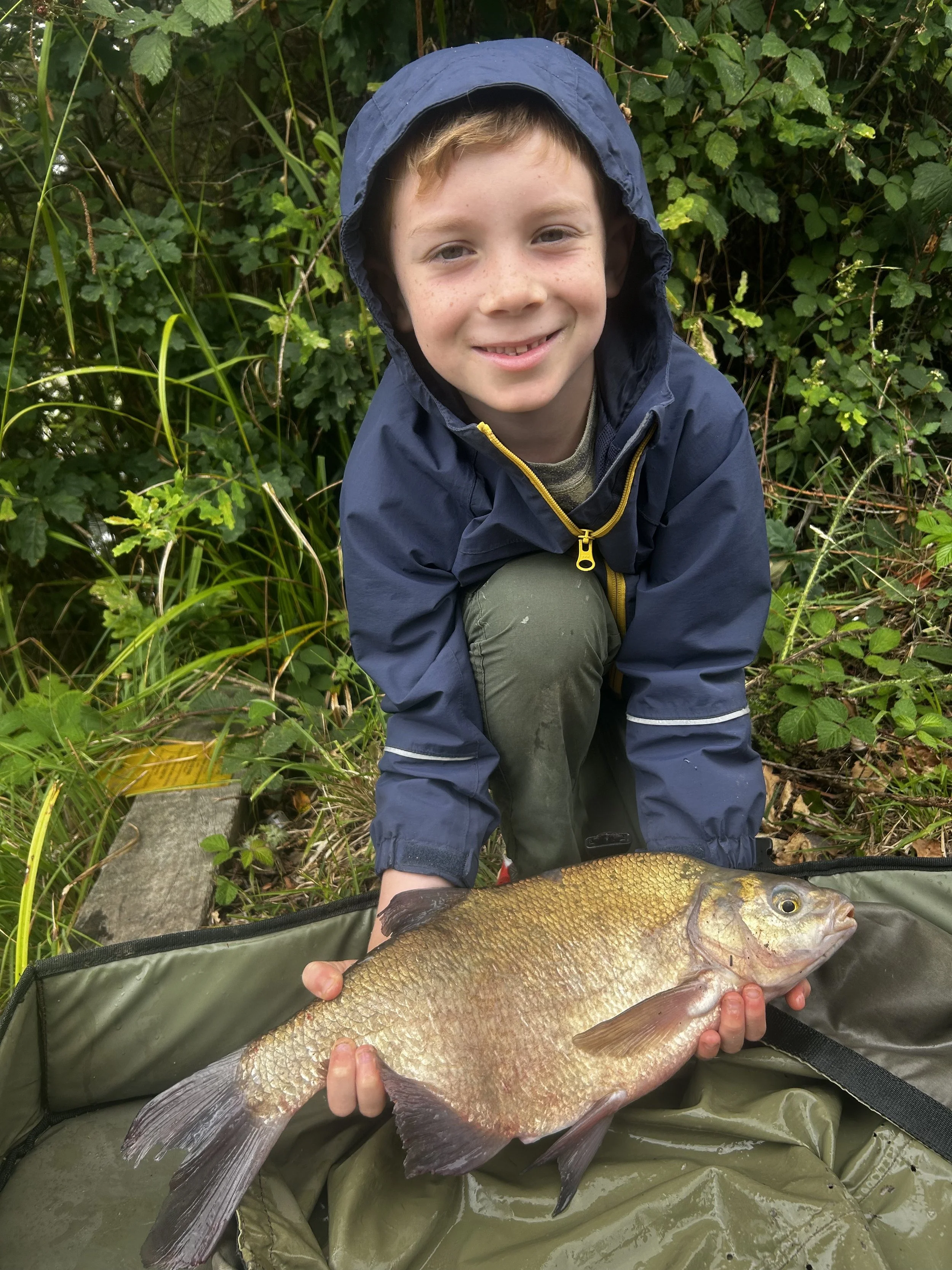 A young boy with a blue rain jacket and green pants kneeling in green foliage, holding a large fish with gold and silver scales and a pointed tail, lying on a green fishing mat.