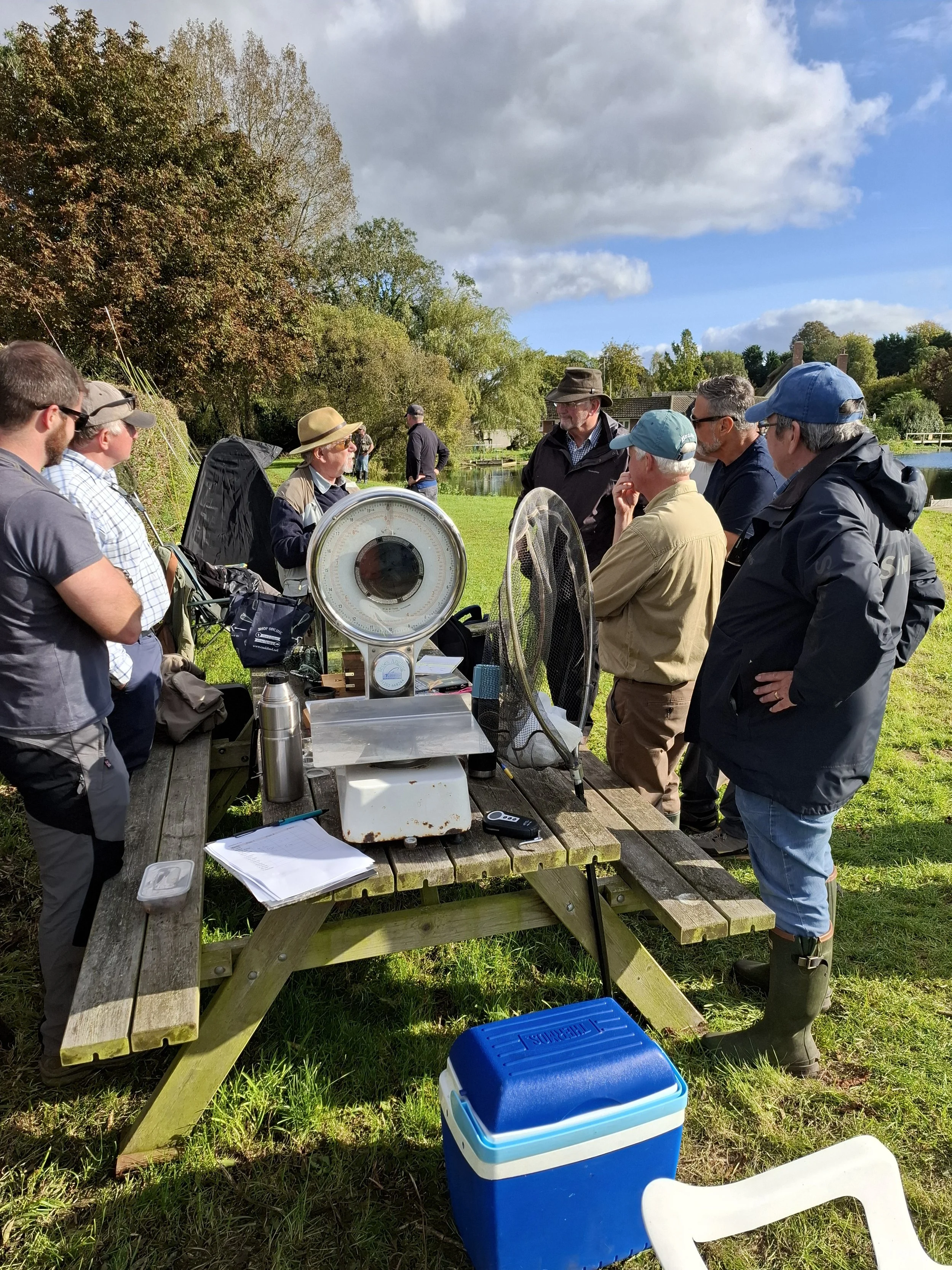 Group of people gathered outdoors around a table with scientific equipment, including weighing scales, in a park with trees and a pond, on a partly cloudy day.