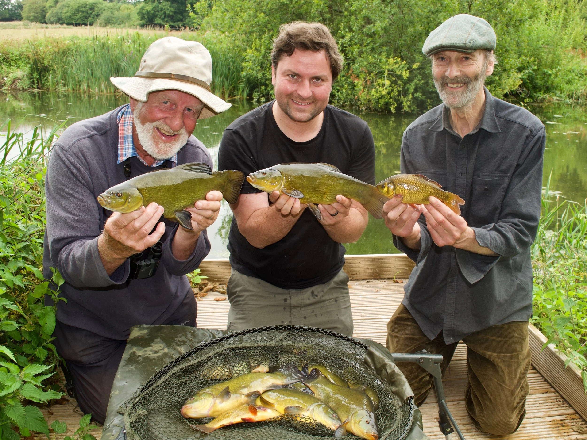 Three men holding large fish by a pond, smiling, outdoors with green trees and plants in the background.
