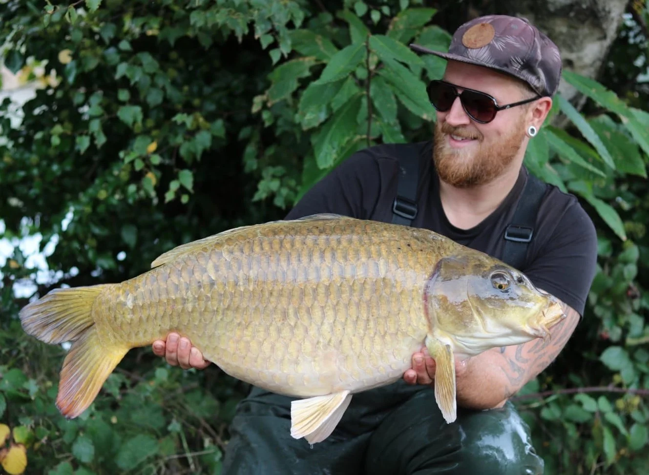 A man with a beard, sunglasses, and a camo cap holding a large fish outdoors.