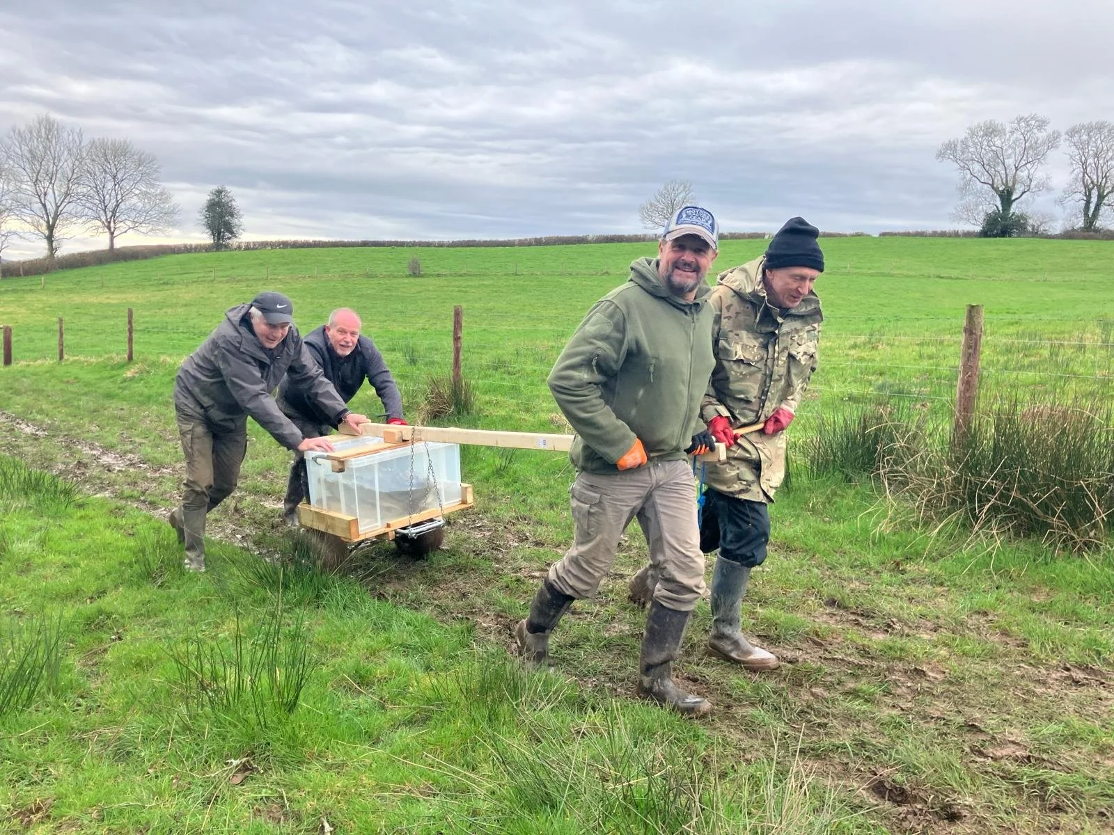 Four men working outdoors in a green field with trees, some dressed in jackets and gloves, moving a makeshift cart with a rectangular container, possibly for scientific or environmental work.