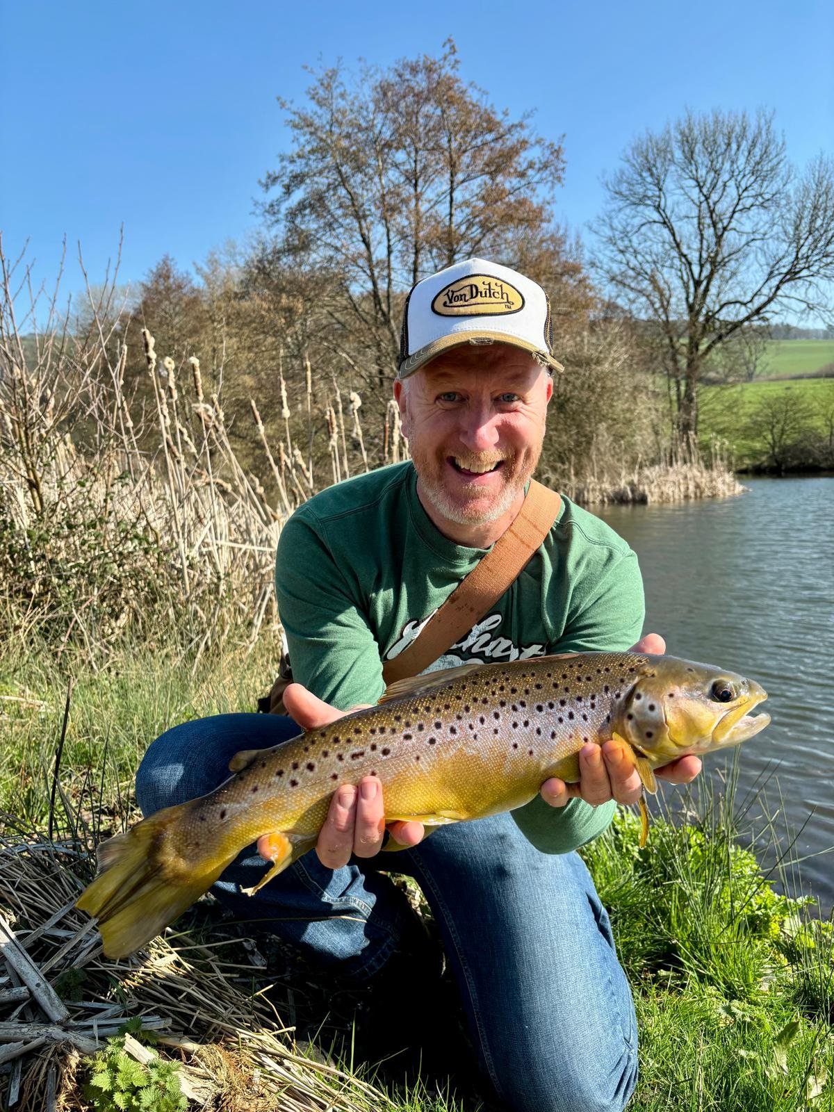 A smiling man wearing a baseball cap and green shirt is kneeling on grass near a river, holding a large brown trout fish he caught.