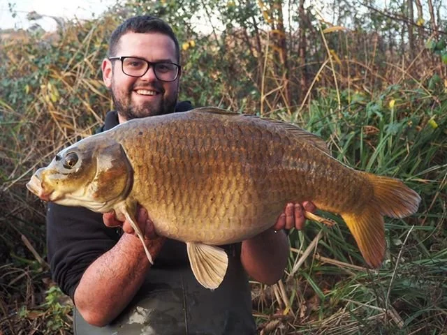 Man with beard and glasses holding a large fish outdoors near reeds and plants.