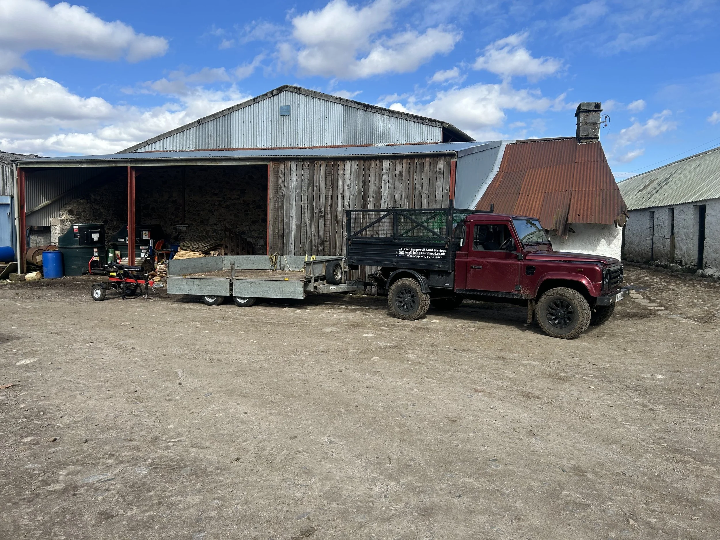 A red pickup truck with a trailer attached, parked on a dirt lot in front of a rustic farm building with metal roof and wood siding, under a partly cloudy sky. Timber transport and tree products. Expert Tree Cutting Aviemore and Inverness.
