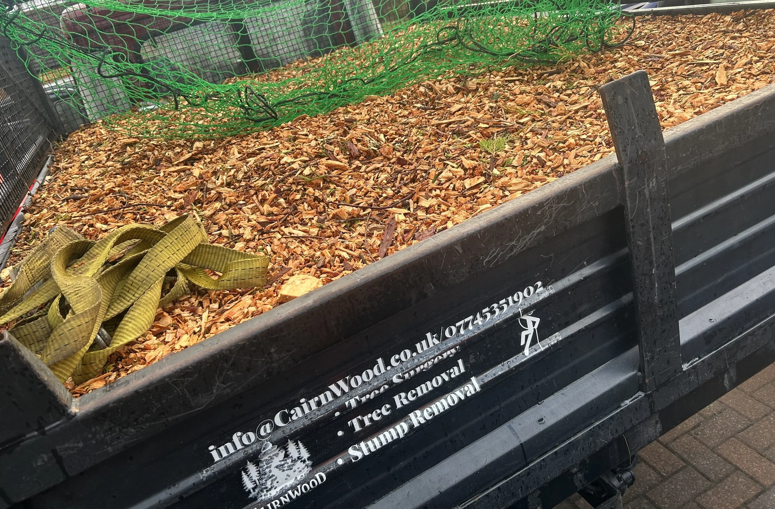 A black trailer filled with wood chippings. There is a metal tool leaning against the trailer and contact information and services, including tree removal and stump removal, written on the side. Local Tree Surgery Located Aviemore.