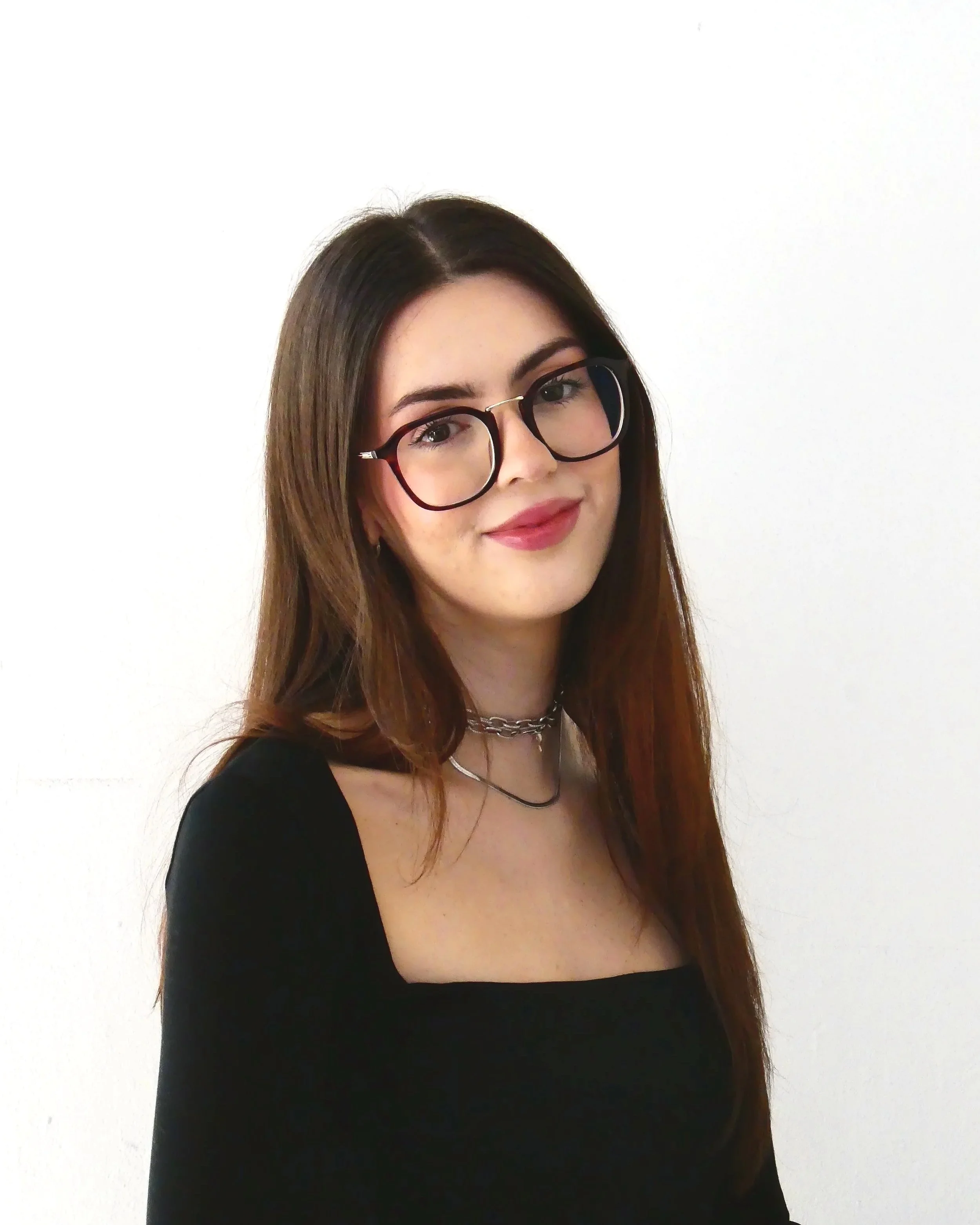 A young woman with long brown hair, wearing black glasses, a black top, and layered necklaces, smiling against a white background.