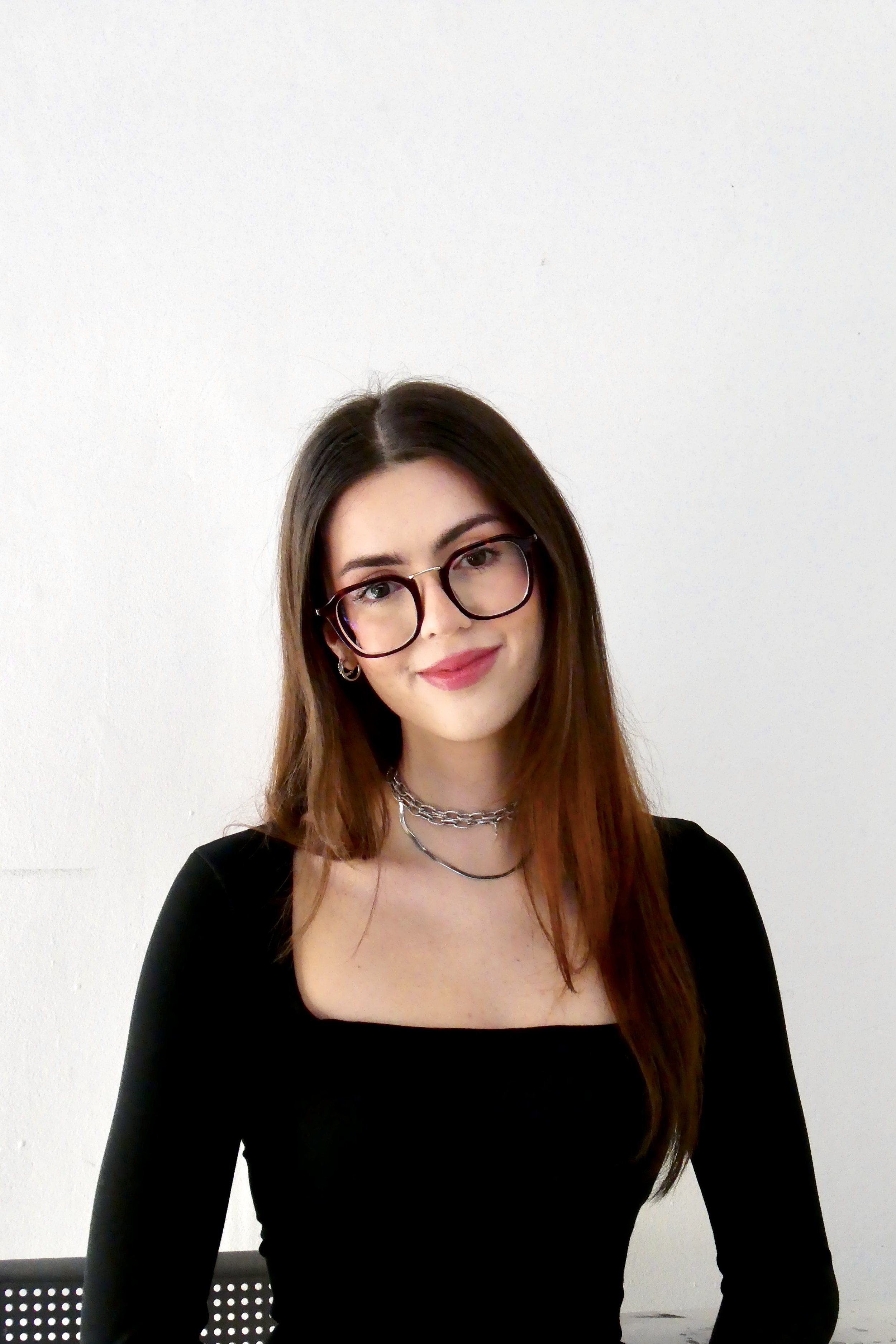 A young woman with long brown hair, wearing glasses and a black top, standing against a white wall.