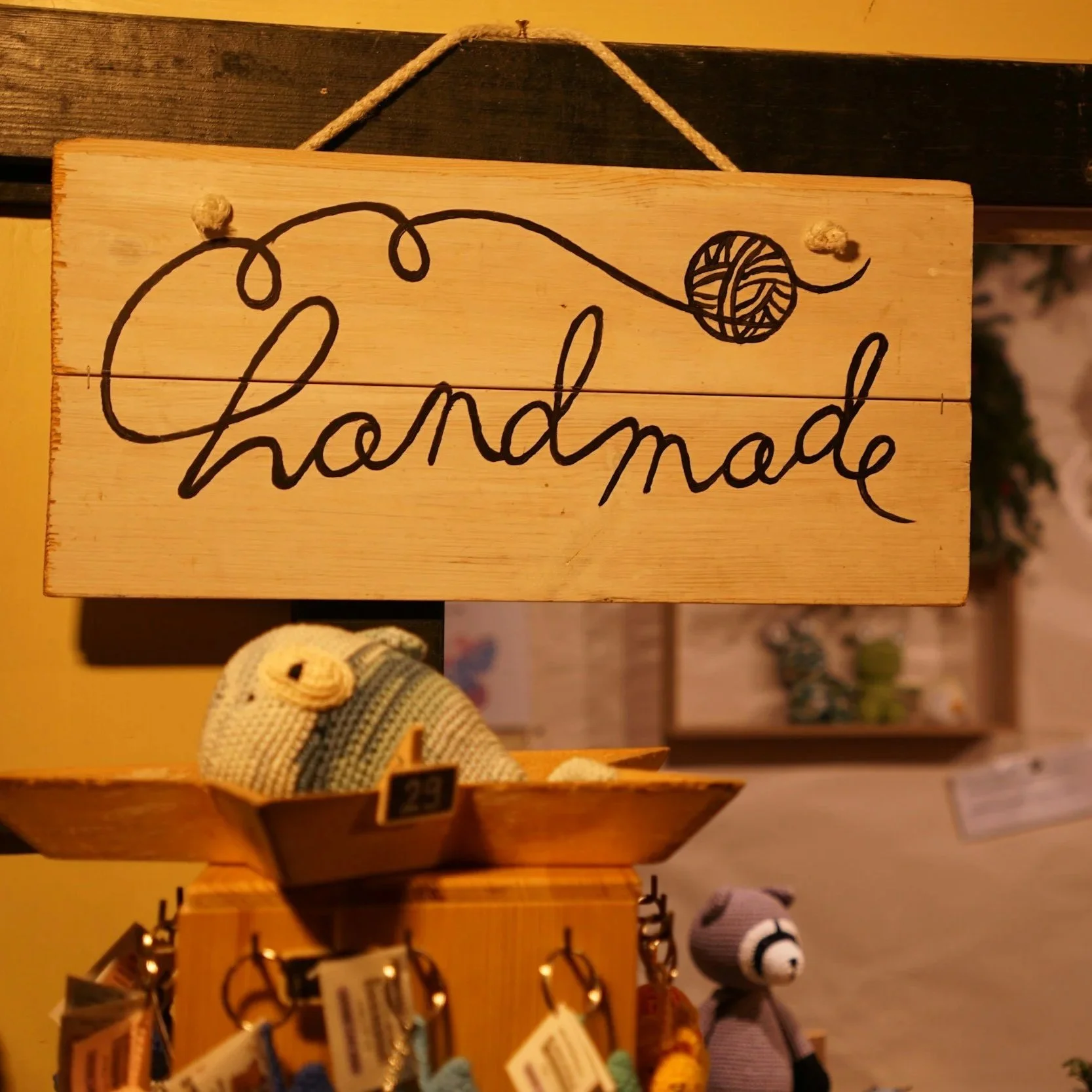Wooden sign with black cursive handwriting that says 'handmade' and an illustration of a ball of yarn with a knitting needle. Below is a small knitted teddy bear in a display.