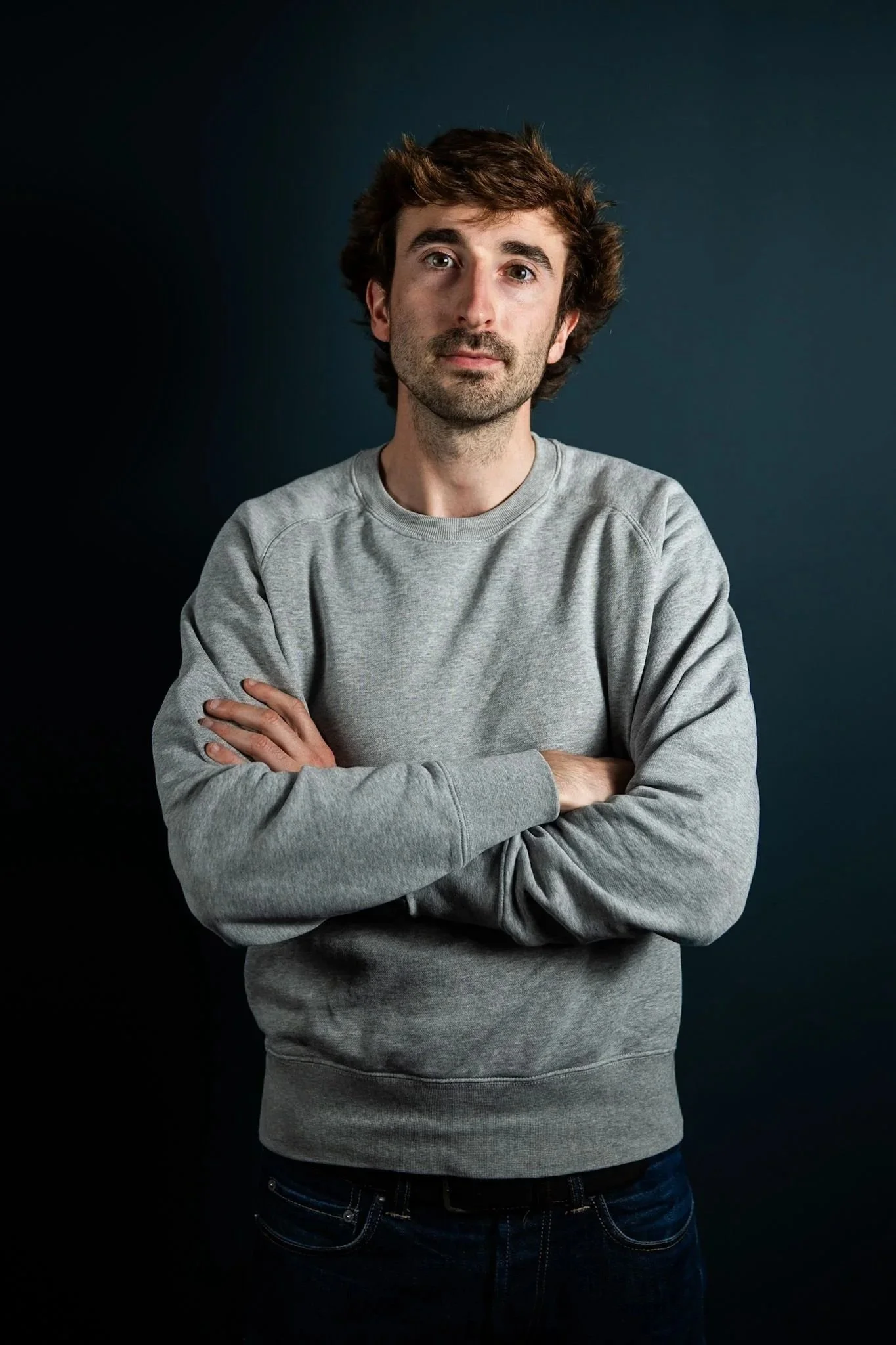 A young man with brown hair and facial hair, wearing a gray sweatshirt, stands with arms crossed against a dark background.