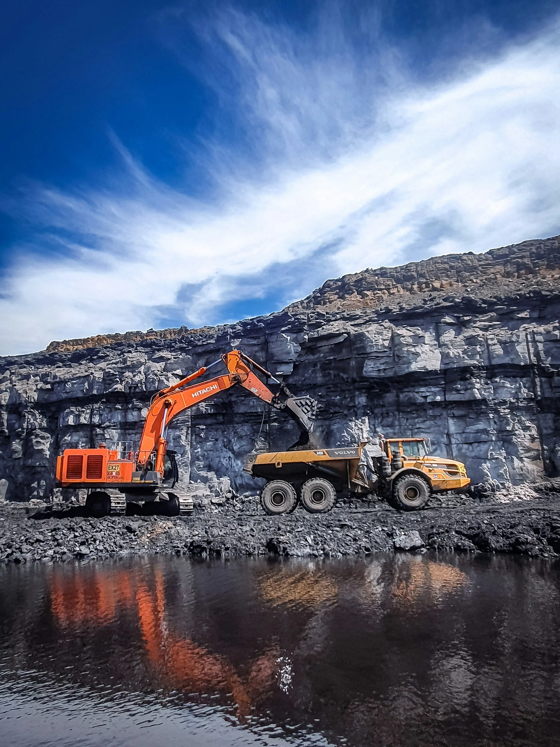 Construction scene with an orange excavator pouring dirt into a dump truck near a rocky cliff and body of water under a blue sky with clouds.
