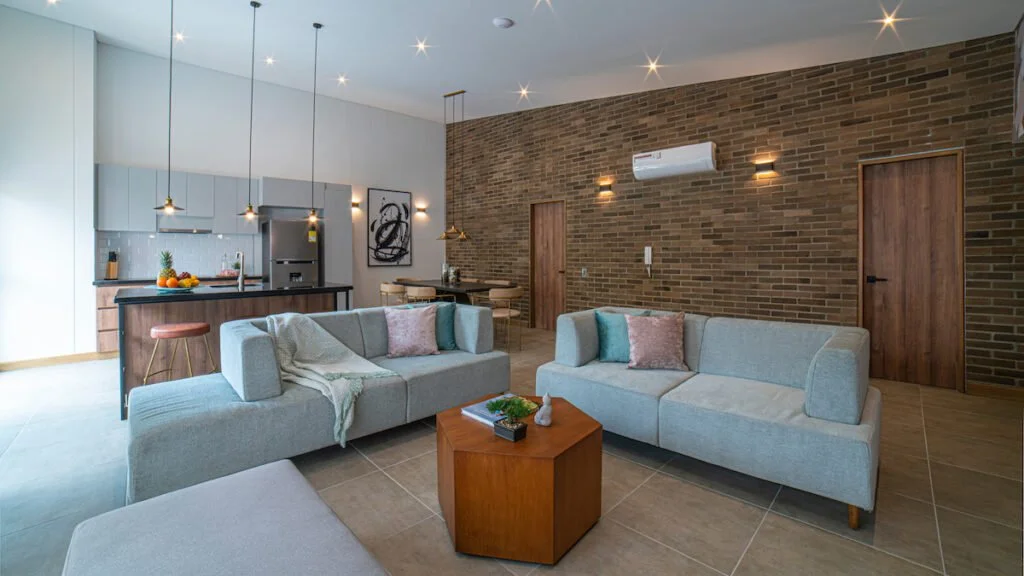 ERE Centenario, unit 302 - Cali, Colombia. 
Modern living room with two light gray sofas, wooden coffee table, and brick accent wall with wall-mounted light fixtures. Open kitchen with island, fruit basket, and dining area in the background.