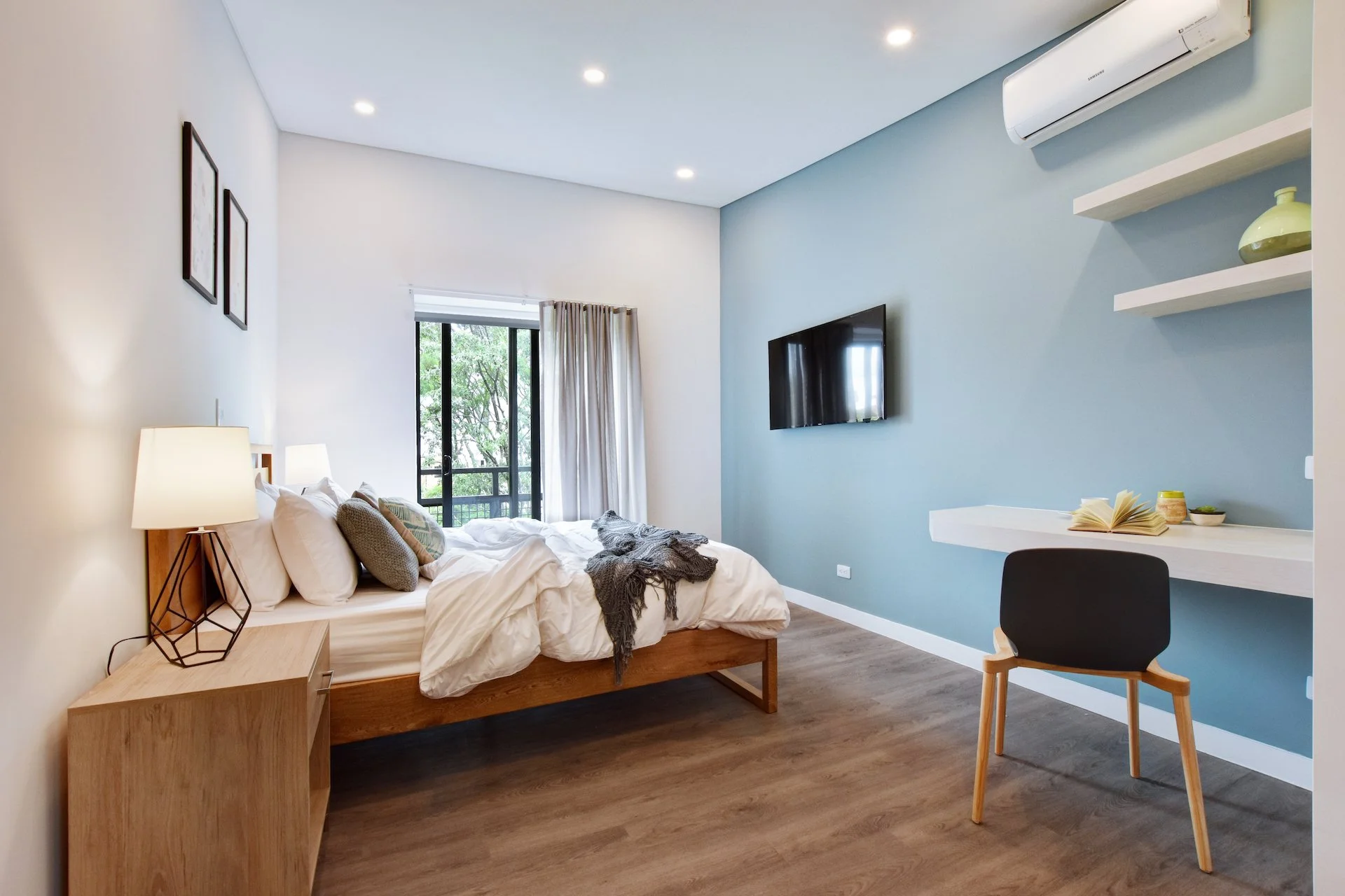 Modern bedroom with a bed, side table, lamp, wall-mounted TV, and a desk with a chair against a blue wall. There is a sliding door leading to a balcony, and the room has wooden flooring and ceiling lights.