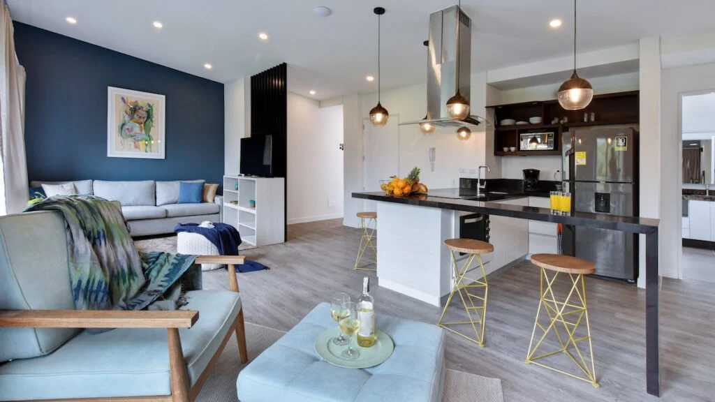 Open-concept living room and kitchen with modern decor, seating area, artwork on the wall, kitchen island with stools, pendant lights, refrigerator, and fruit on the counter.