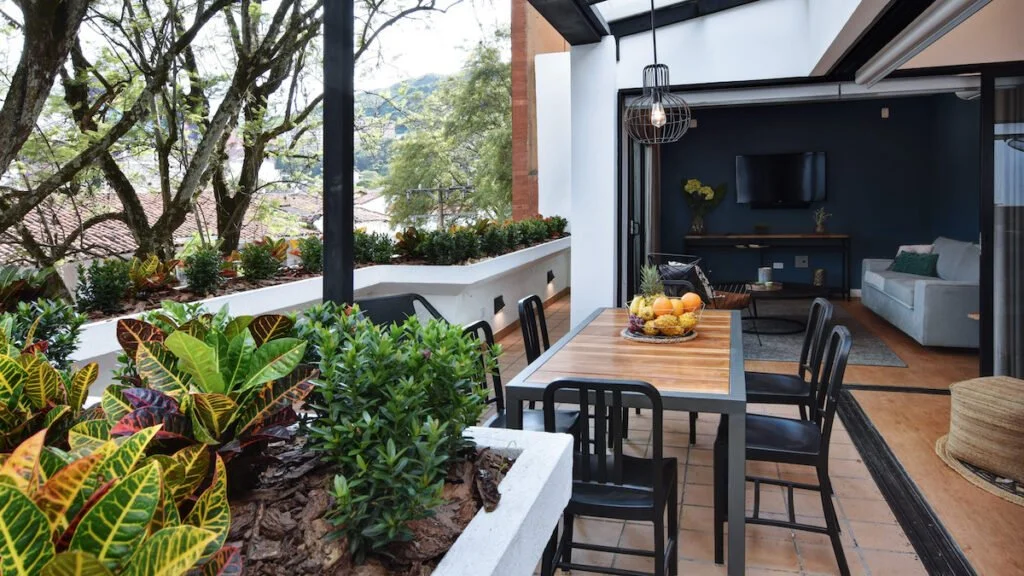 Outdoor patio with a wooden dining table and six black chairs, surrounded by potted plants and greenery, leading into a modern indoor living room with a sofa, television, and cozy decor.