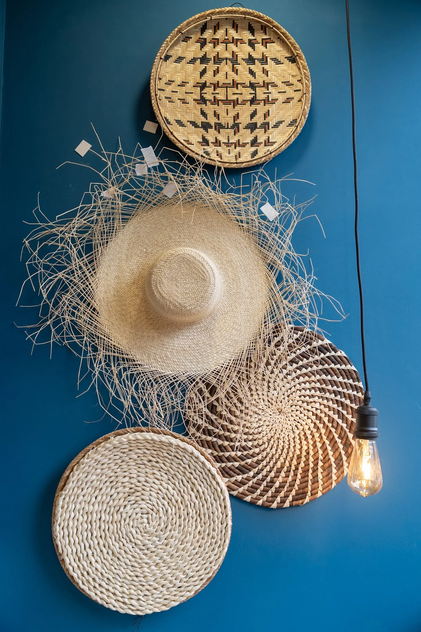 Decorative woven trays and a straw sunhat hung on a blue wall with a hanging lightbulb.
