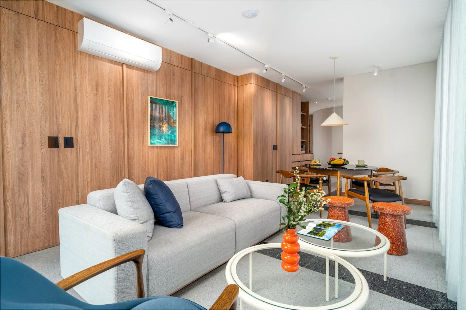 Living room with a white sofa, blue cushions, wooden wall paneling, and a round coffee table with a red vase holding white flowers. In the background is a dining area with a black table, wooden chairs, and a hanging white pendant light.