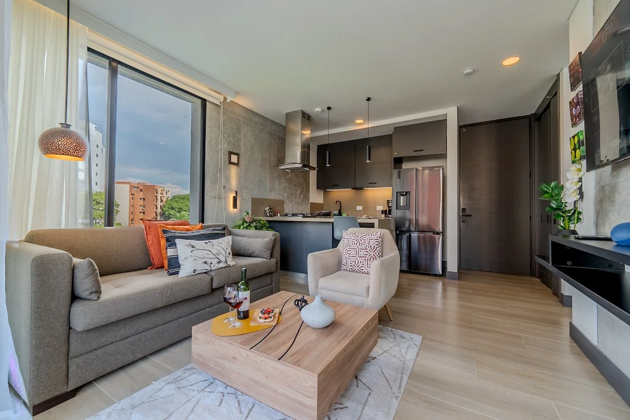 Modern open-concept living room and kitchen with large window, beige sofa, white armchair, wooden coffee table, and stainless steel refrigerator.