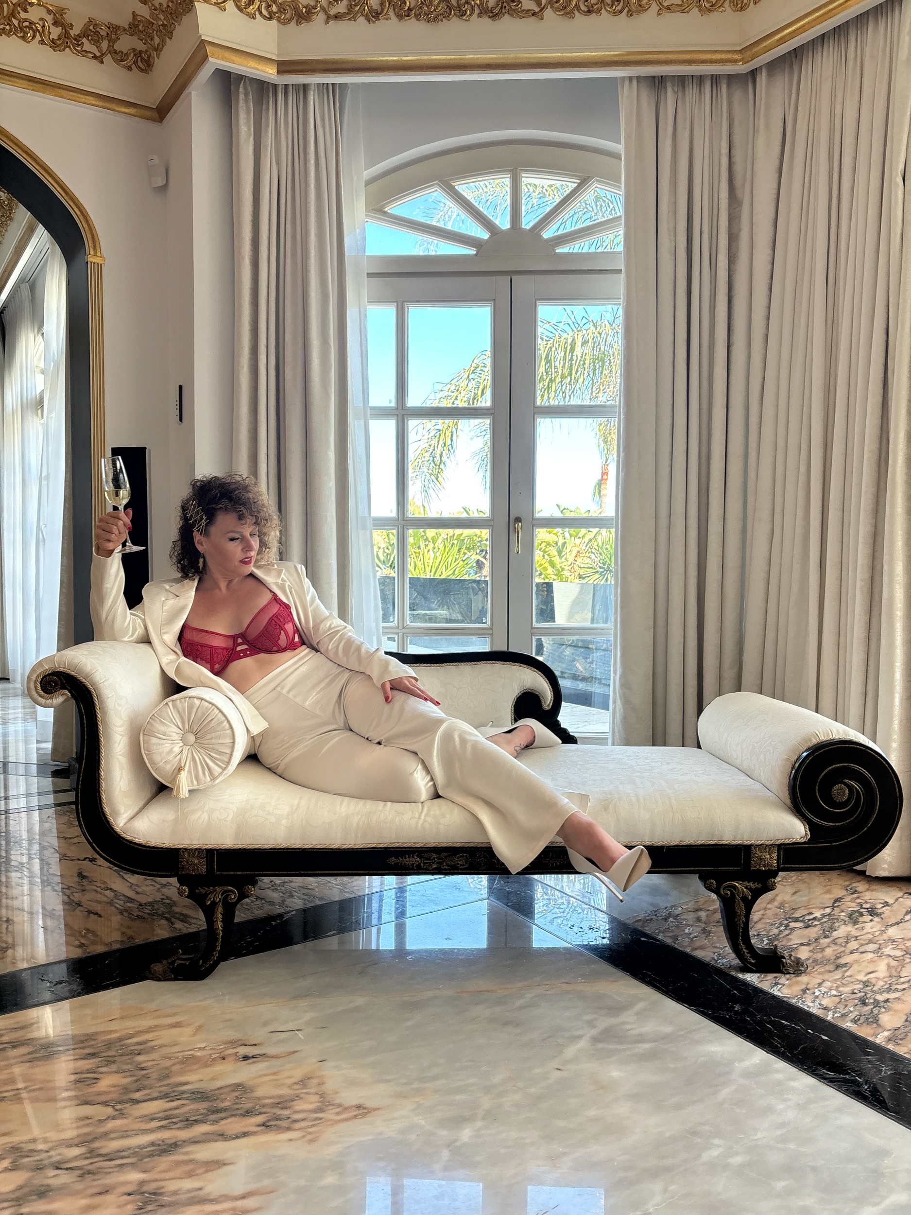 A woman in red lingerie sitting on a white sofa in a luxurious room with ornate gold decorations, marble flooring, and large windows with curtains.  An international companion, muse, and guide in sensuality and intimacy. Available worldwide.