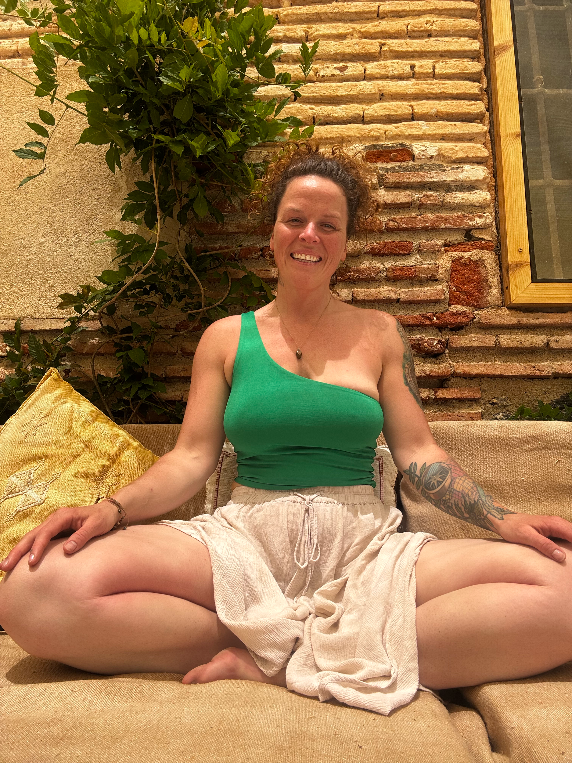 A woman with curly brown hair and tattoos on her arm is sitting on a yellow lounge chair with beige cushions outdoors, smiling at the camera.  An international companion, muse, and guide in sensuality and intimacy. Available worldwide.