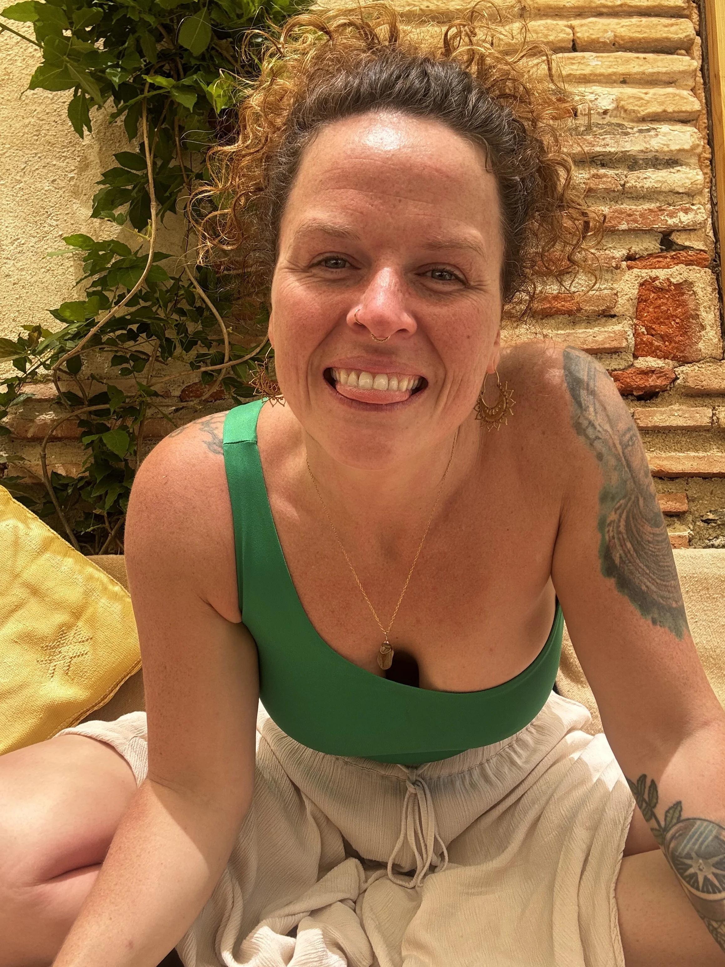 A woman with curly hair and tattoos wearing a green top and beige shorts, smiling. She is sitting against a brick wall with greenery on the left side.  An international companion, muse, and guide in sensuality and intimacy. Available worldwide.