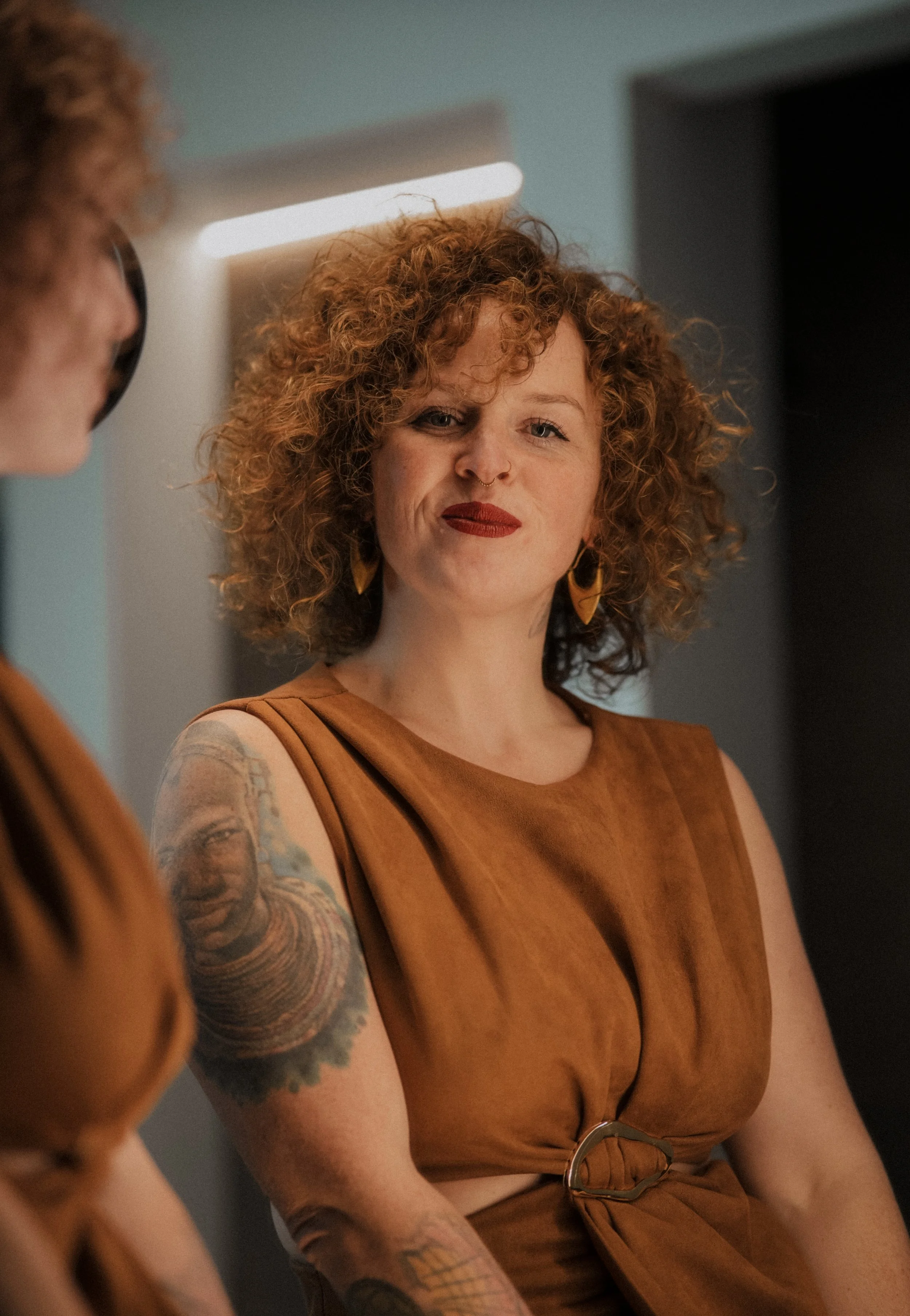 A woman with curly red hair, tattooed arms, wearing a rust-colored dress, sitting in a room with blue walls and a doorframe.  An international companion, muse, and guide in sensuality and intimacy. Available worldwide.