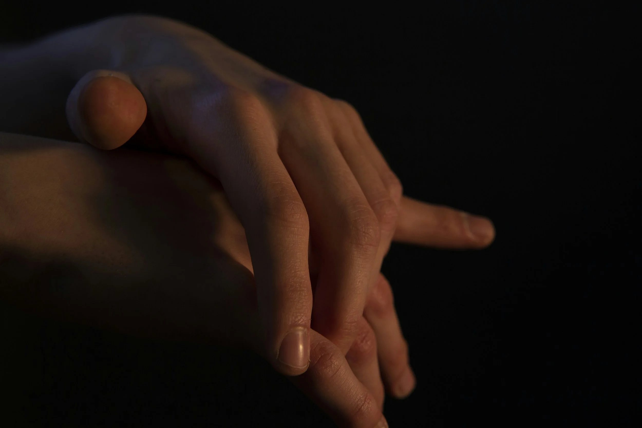 Close-up of a person's hand gently holding another hand against a dark background, with soft lighting highlighting the skin texture.  An international companion, muse, and guide in sensuality and intimacy. Available worldwide.