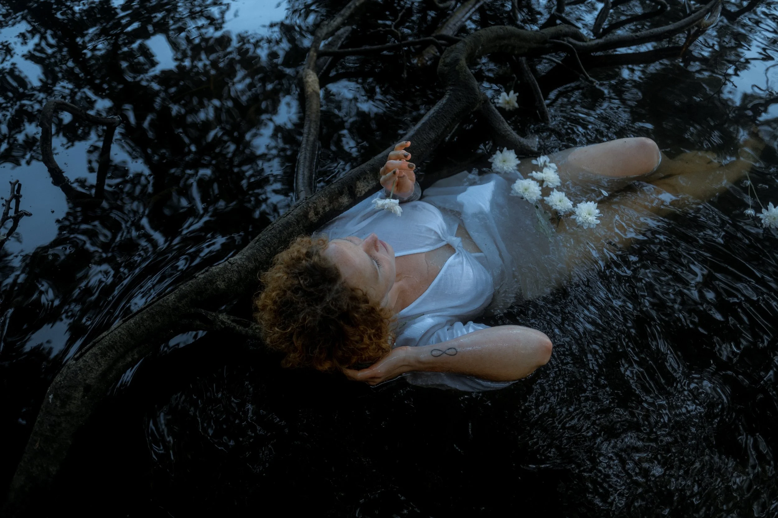 A person with curly hair and a tattoo on their arm is lying in water surrounded by dark tree branches, holding a white flower in their hand.  An international companion, muse, and guide in sensuality and intimacy. Available worldwide.