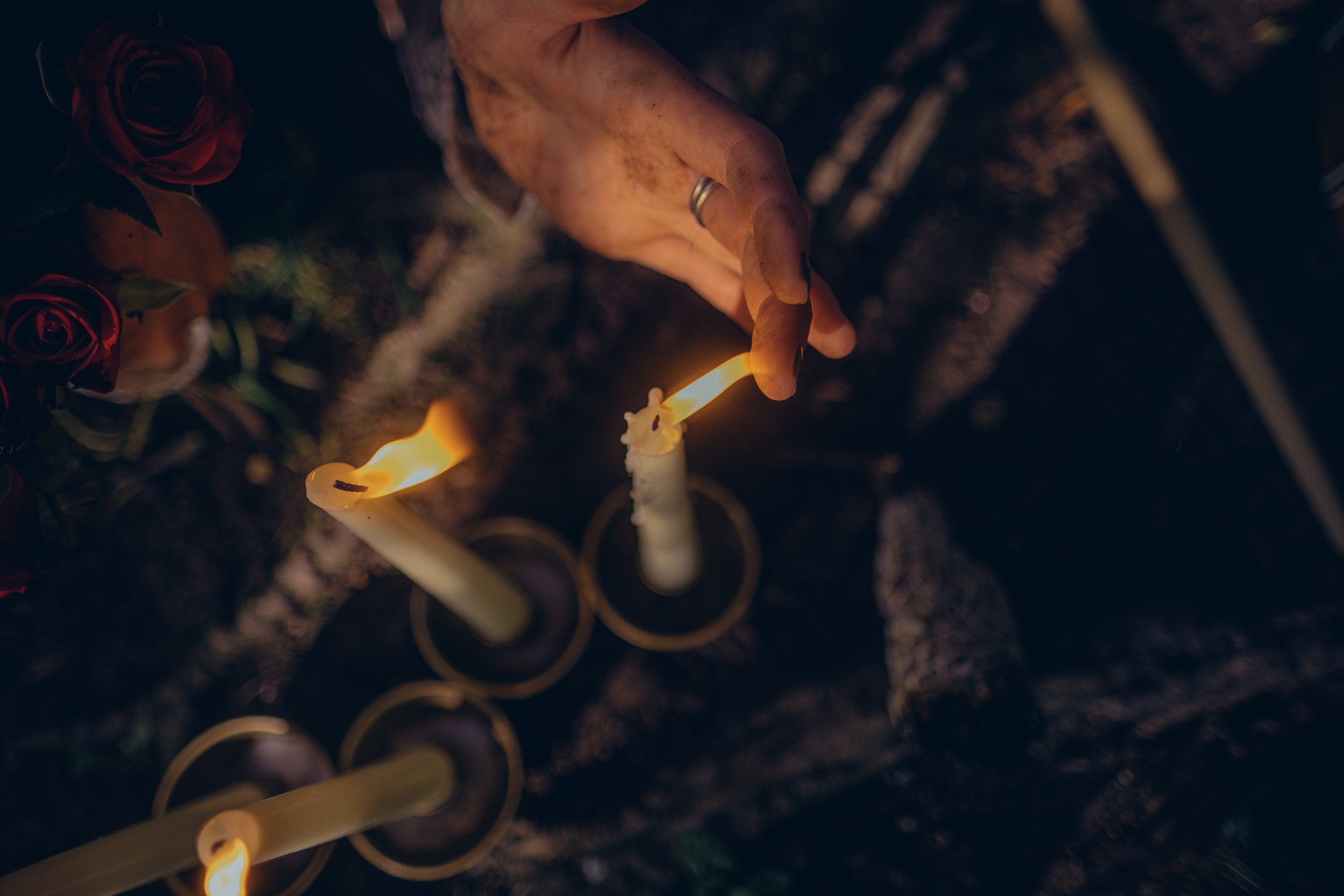 A hand with a wedding band is holding a lit candle, with red roses and more candles in the background, in a dark setting.  An international companion, muse, and guide in sensuality and intimacy. Available worldwide.