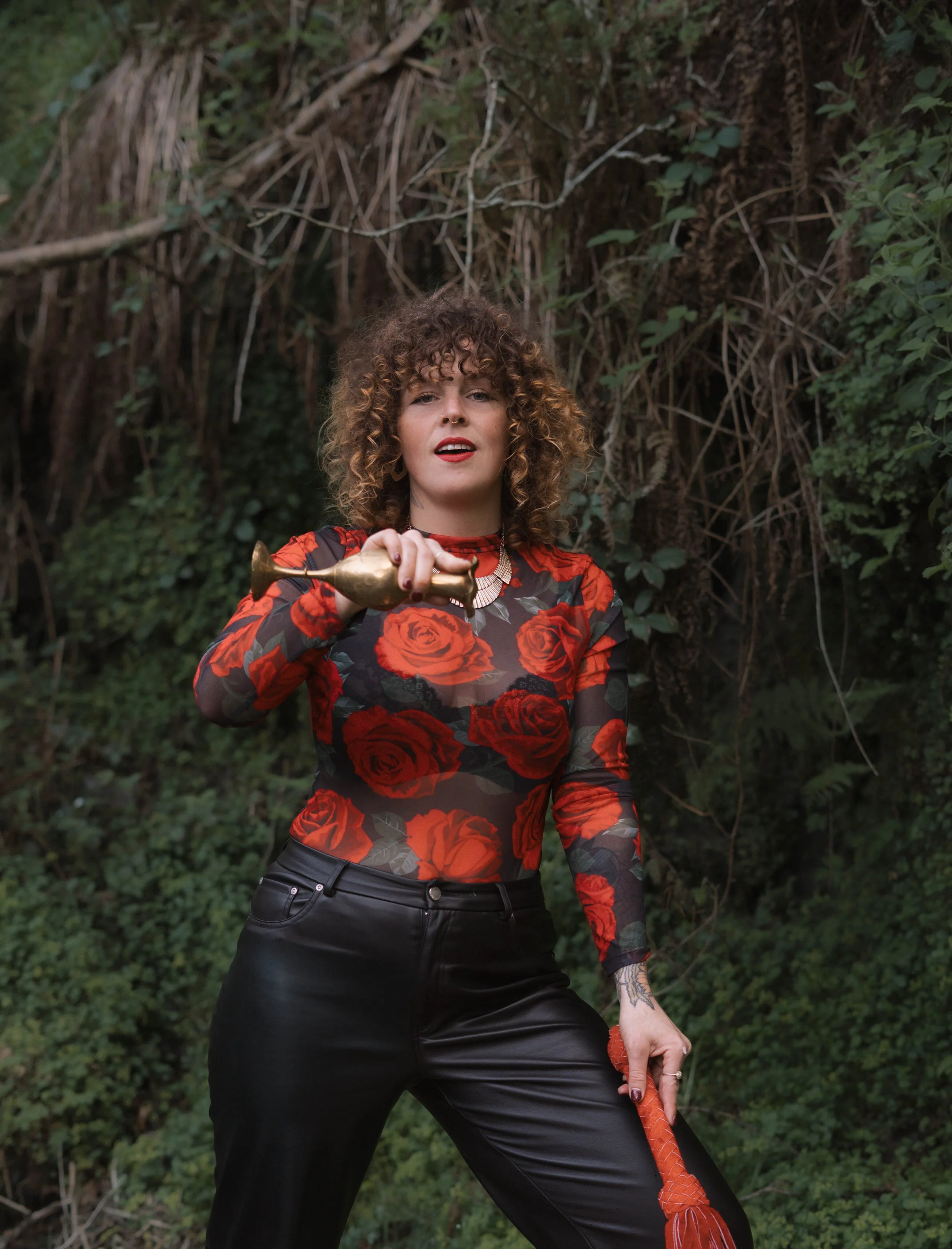 A woman with curly hair and a septum piercing, wearing a floral blouse with red roses and black leather pants, is standing outdoors in front of a leafy.  An international companion, muse, and guide in sensuality and intimacy. Available worldwide.