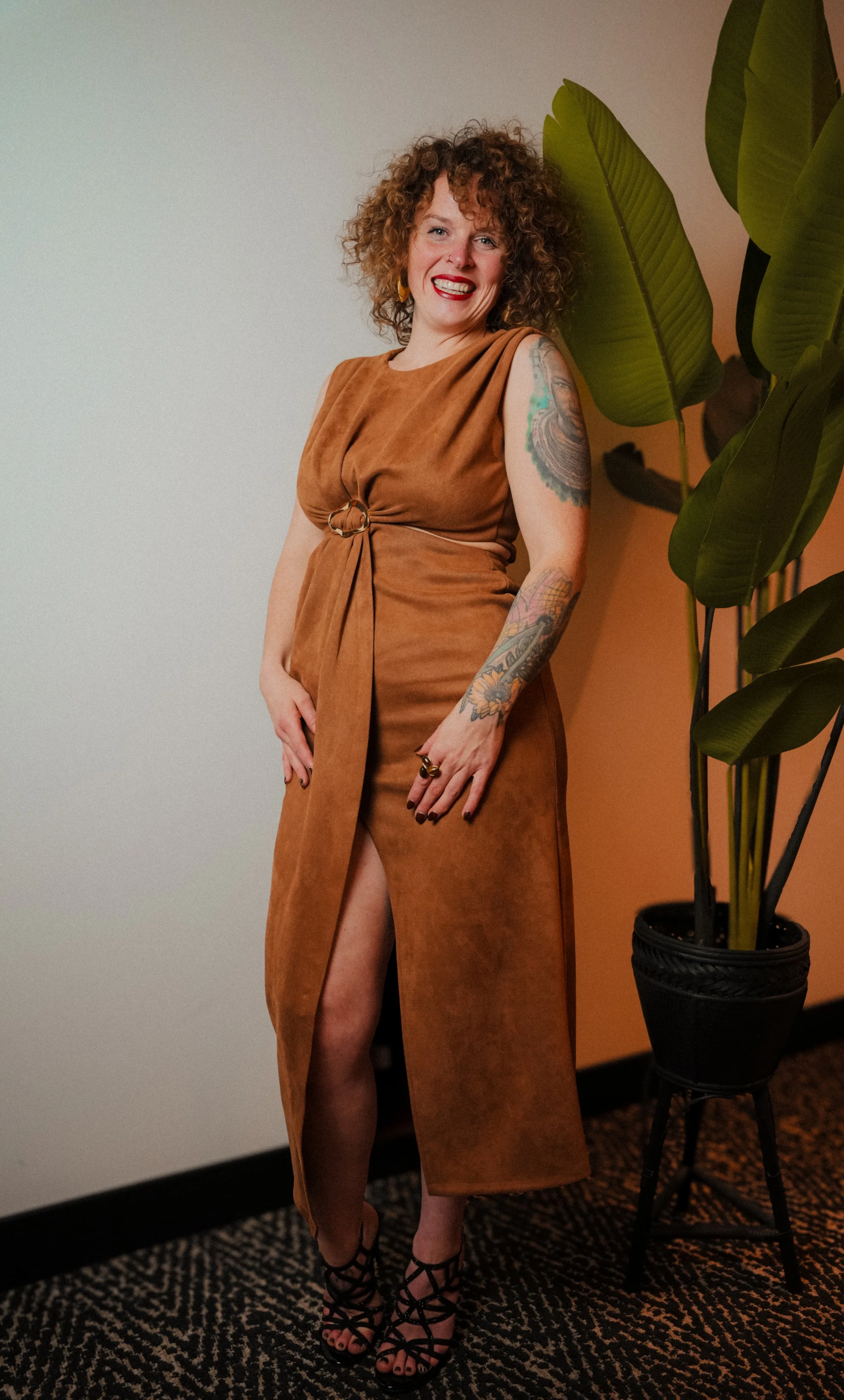 A woman with curly red hair wearing a brown dress with a high slit, black strappy heels, and tattoos on her arms, standing next to a large green plant and smiling.  An international companion, muse, and guide in sensuality and intimacy.