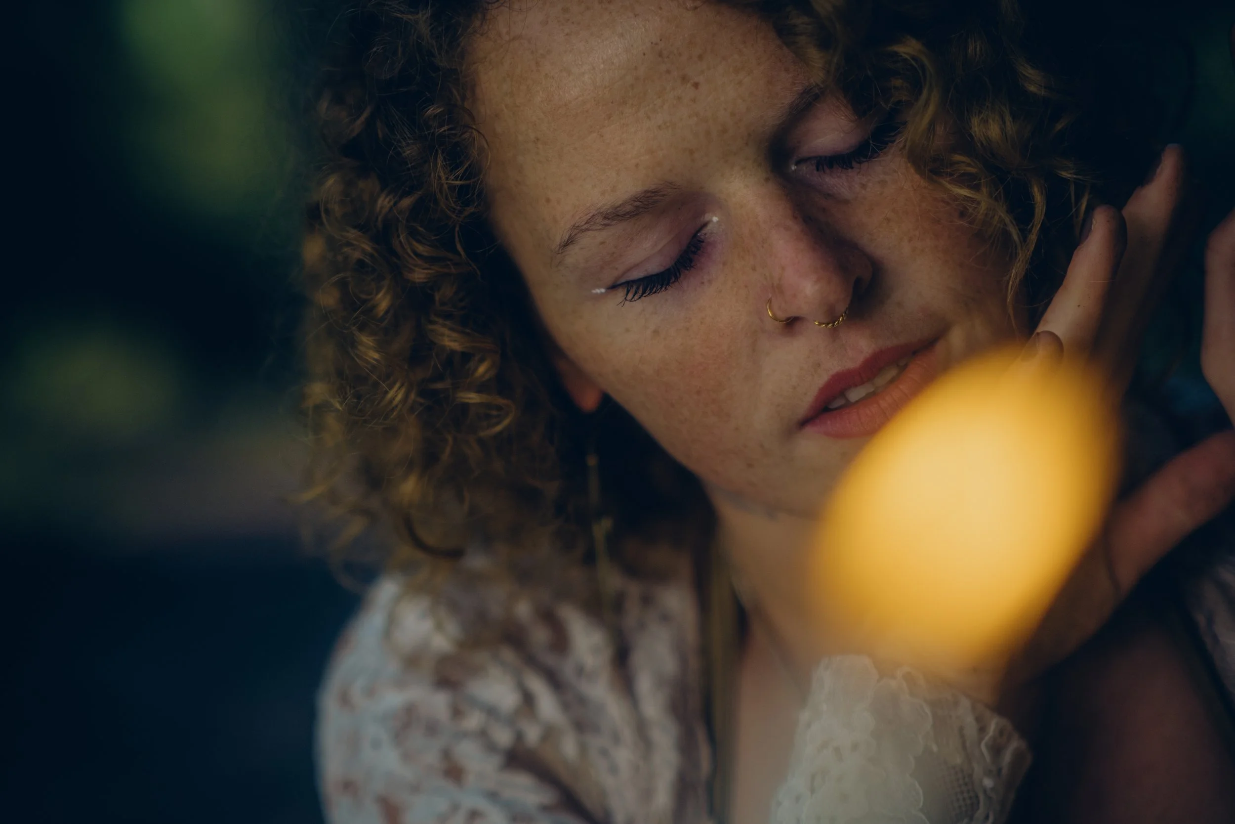 Close-up of a young woman with curly hair, freckles, closed eyes, and a nose ring, smiling gently, with a blurred yellow object in the foreground.  An international companion, muse, and guide in sensuality and intimacy. Available worldwide.