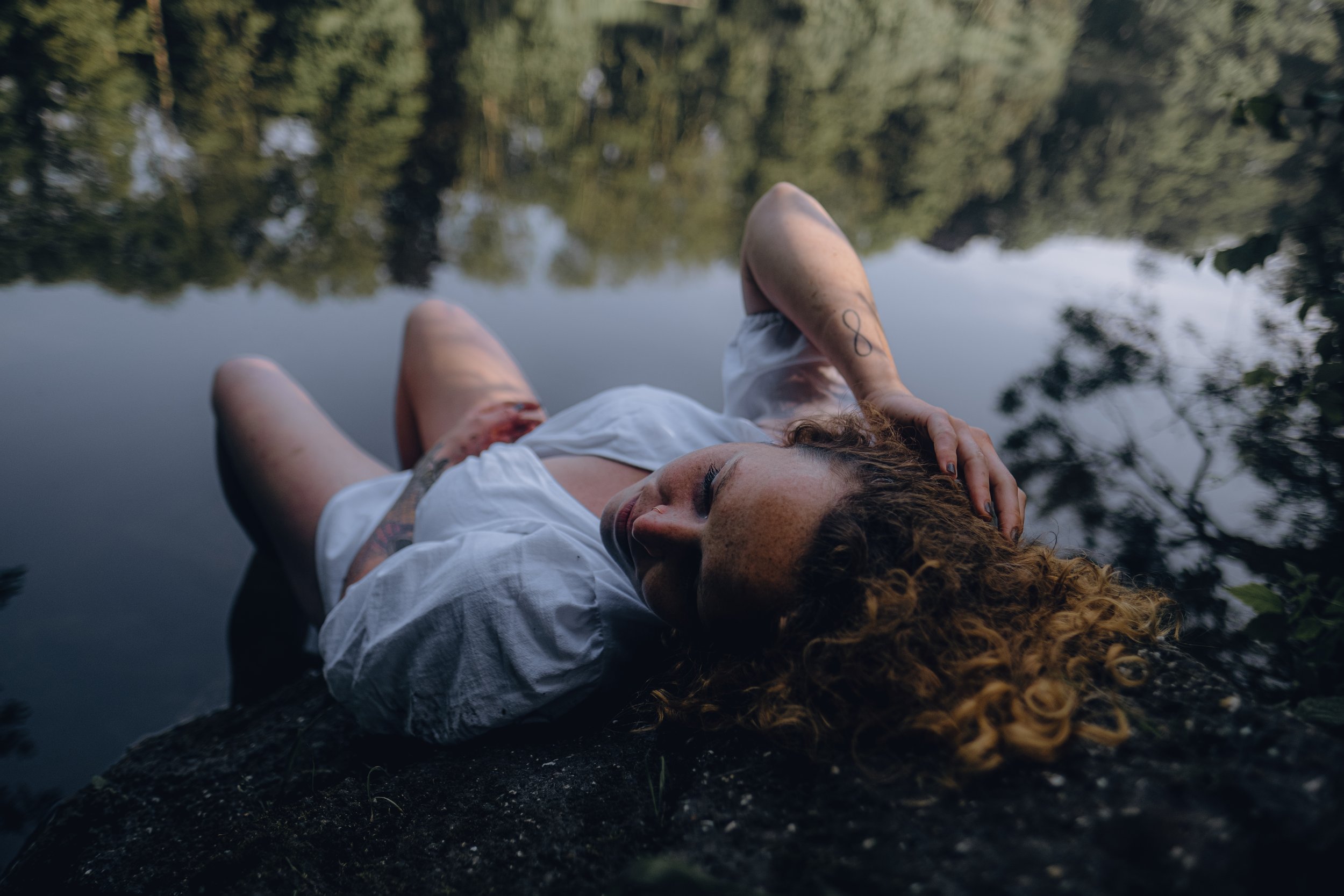 A woman with curly red hair and a tattoo on her arm is lying near a body of water, with trees and their reflections visible on the water's surface.  An international companion, muse, and guide in sensuality and intimacy. Available worldwide.