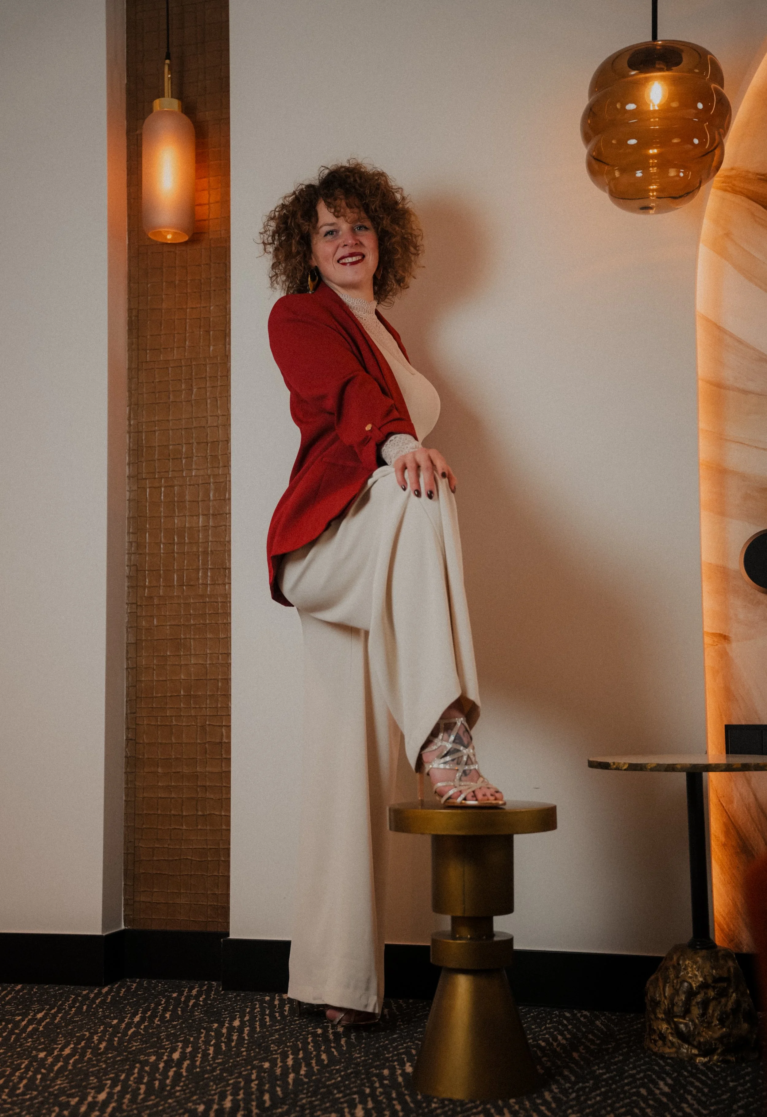 Woman with curly hair wearing a red blazer and white dress standing on a round pedestal in a modern interior with warm lighting.  An international companion, muse, and guide in sensuality and intimacy. Available worldwide.
