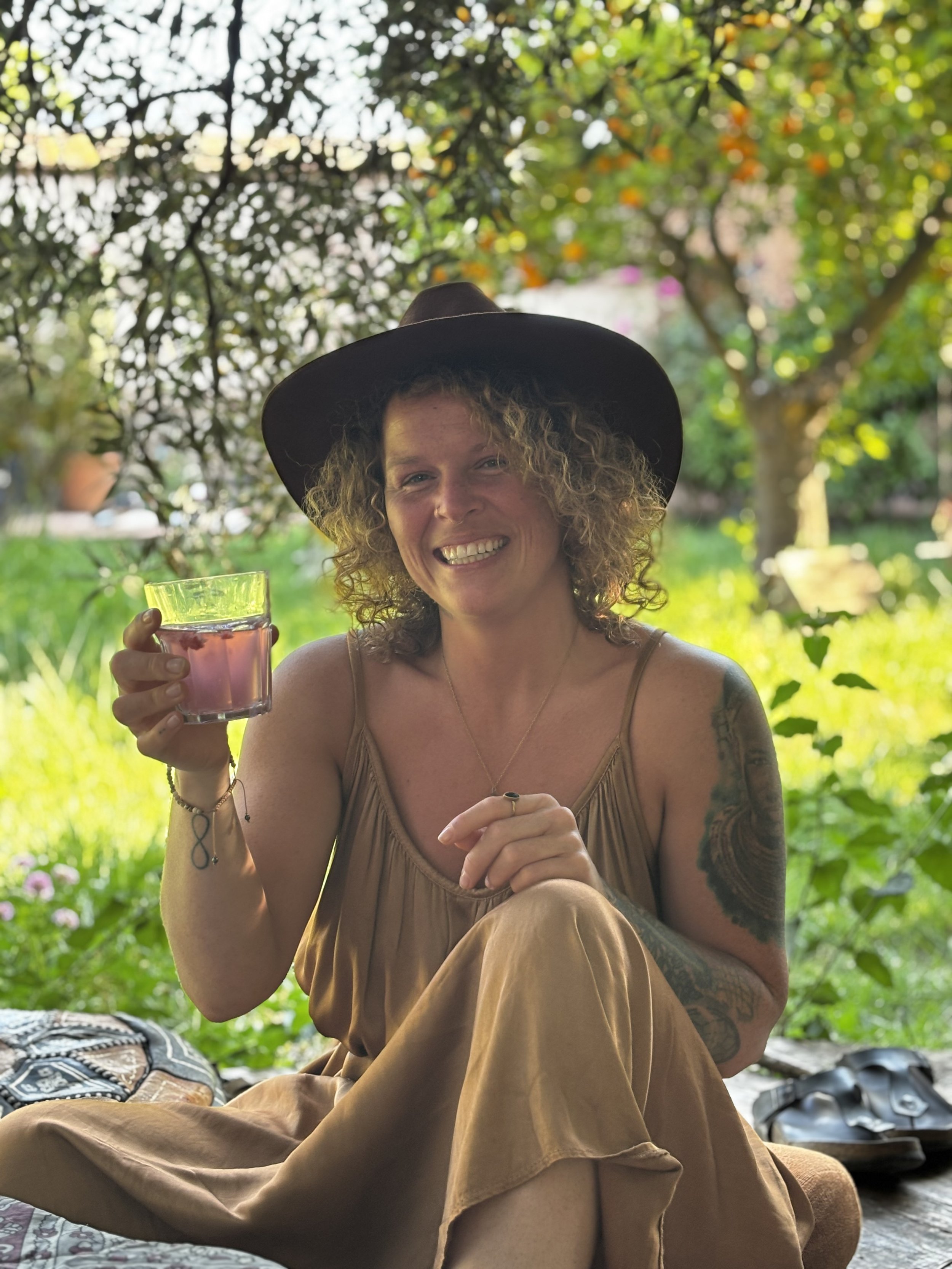 A woman with curly hair wearing a large hat, sitting outdoors under trees, holding a glass of pink drink, smiling.  An international companion, muse, and guide in sensuality and intimacy. Available worldwide.