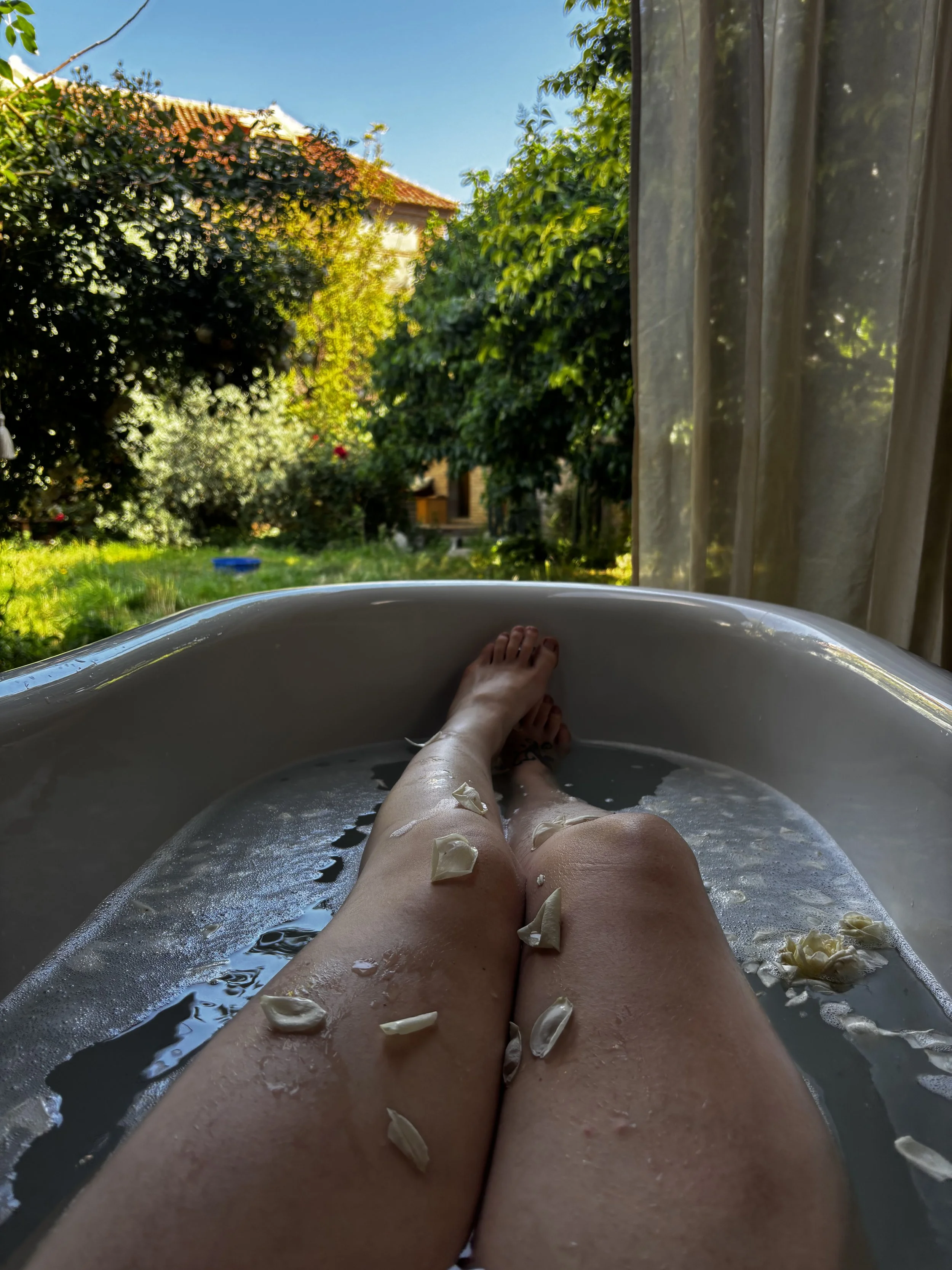 Person soaking in a bathtub with flower petals on legs, looking out a window showing a lush garden and blue sky.  An international companion, muse, and guide in sensuality and intimacy. Available worldwide.