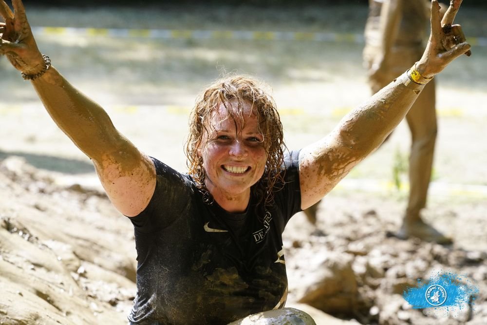 A woman with wet, curly hair smiling and raising her arms after participating in a muddy outdoor event.  An international companion, muse, and guide in sensuality and intimacy. Available worldwide.