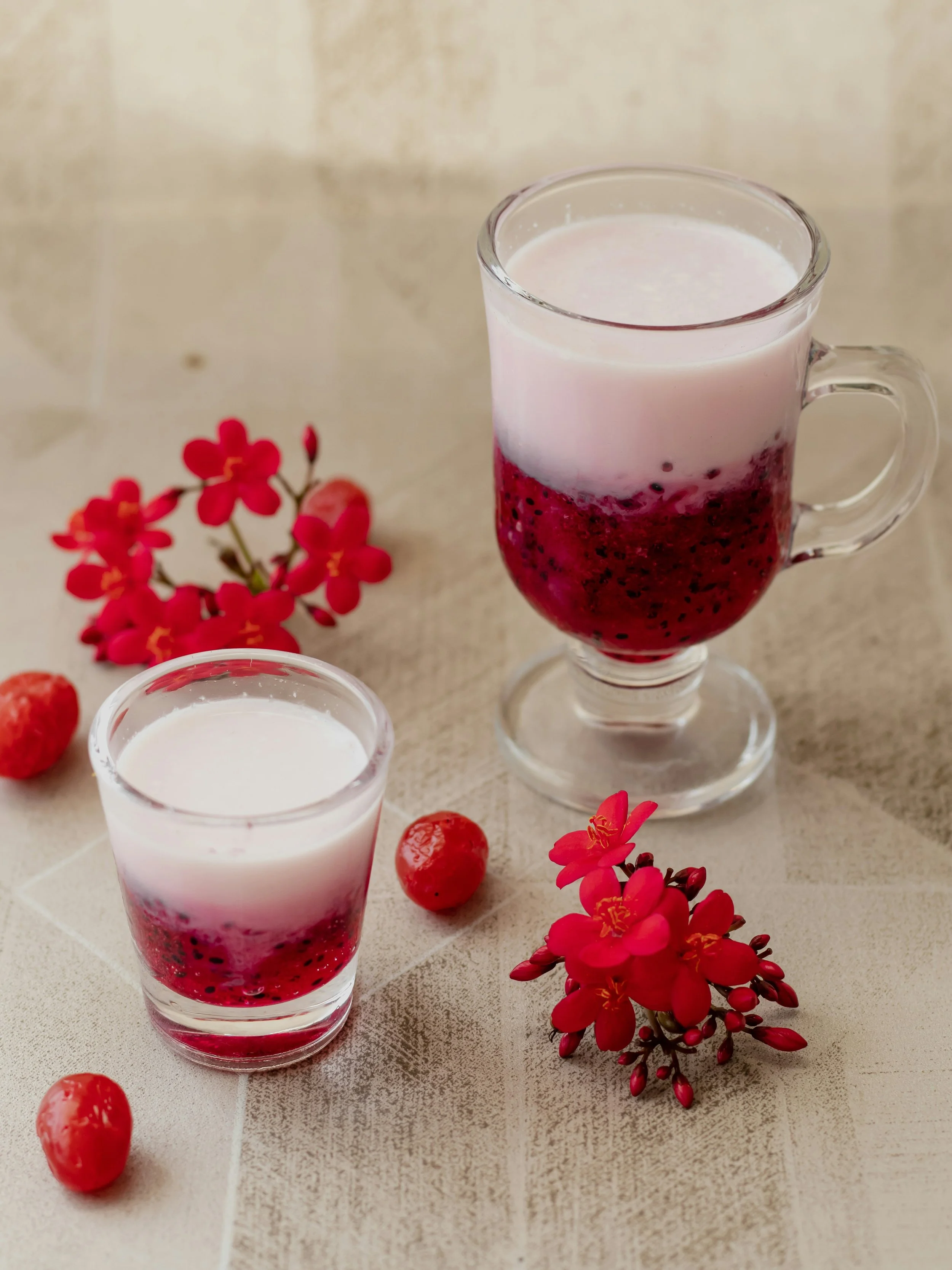 A glass mug with a layered berry and cream drink, surrounded by red berries and pink flowers on a beige surface.  An international companion, muse, and guide in sensuality and intimacy. Available worldwide.