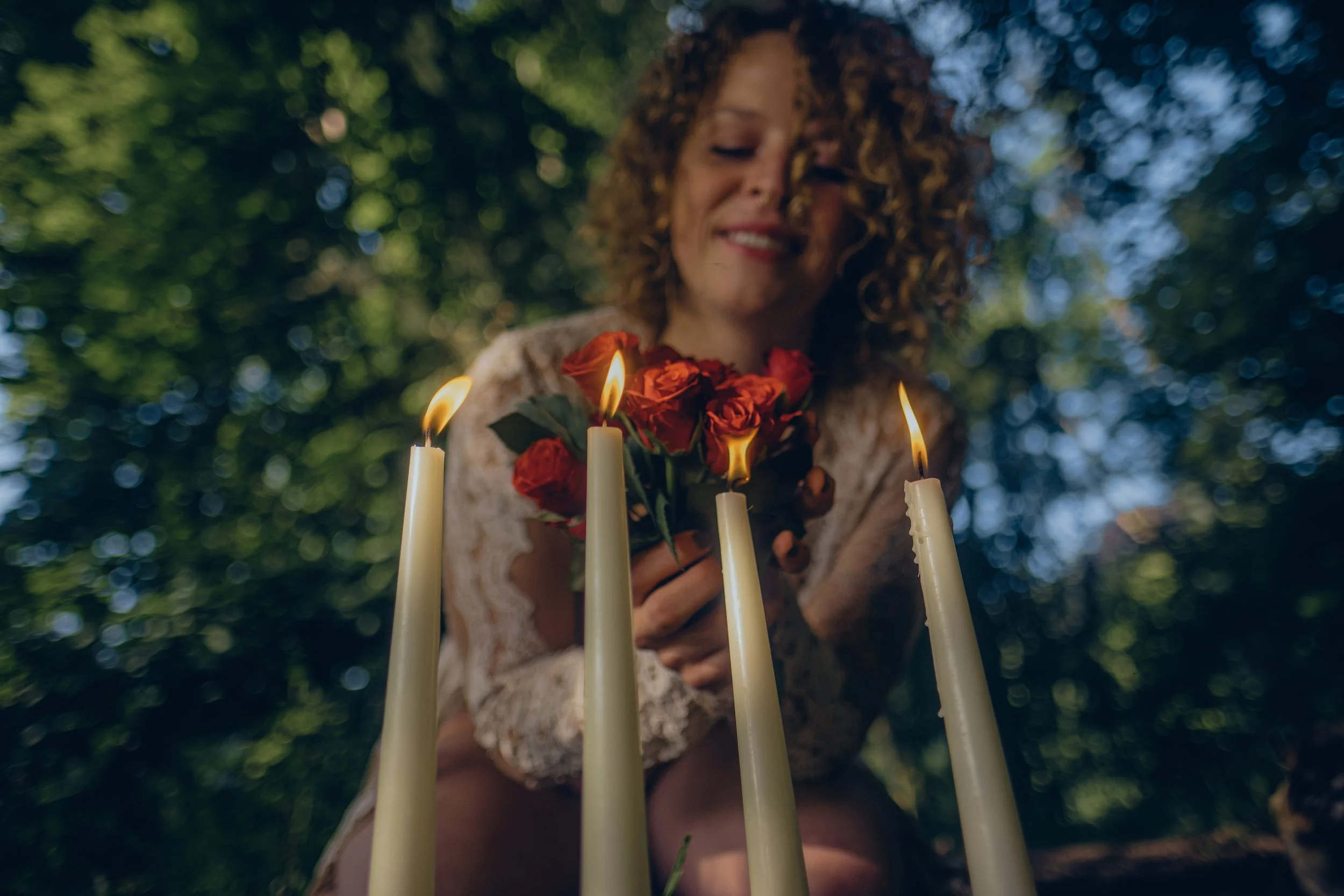 A woman with curly hair holding a bouquet of red roses and standing behind five lit white candles outdoors surrounded by green trees.  An international companion, muse, and guide in sensuality and intimacy. Available worldwide.