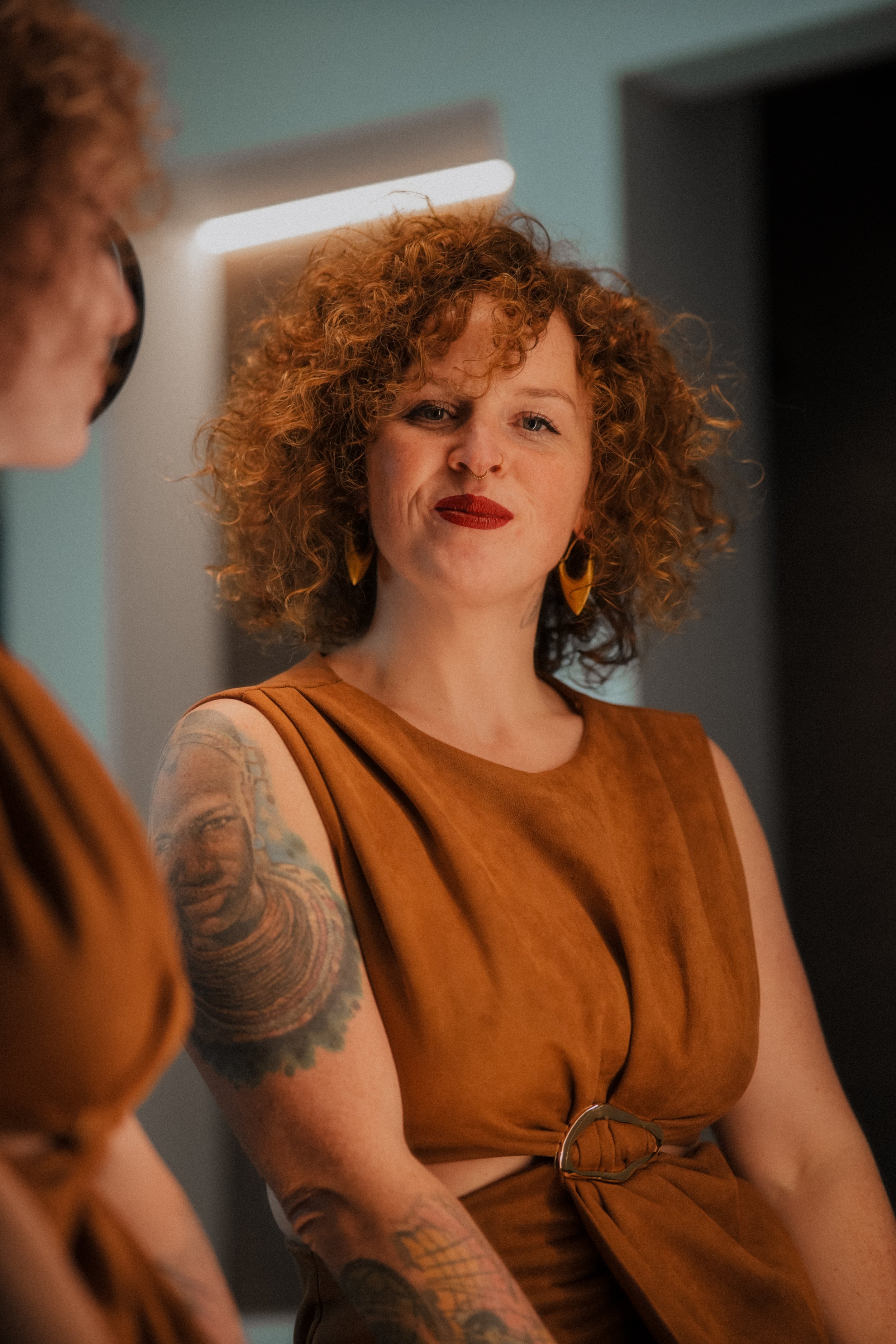 A woman with curly red hair and tattoos on her arm, wearing a brown sleeveless dress with a belt, sitting and looking at her reflection in a mirror. An international companion, muse, and guide in sensuality and intimacy. Available worldwide.