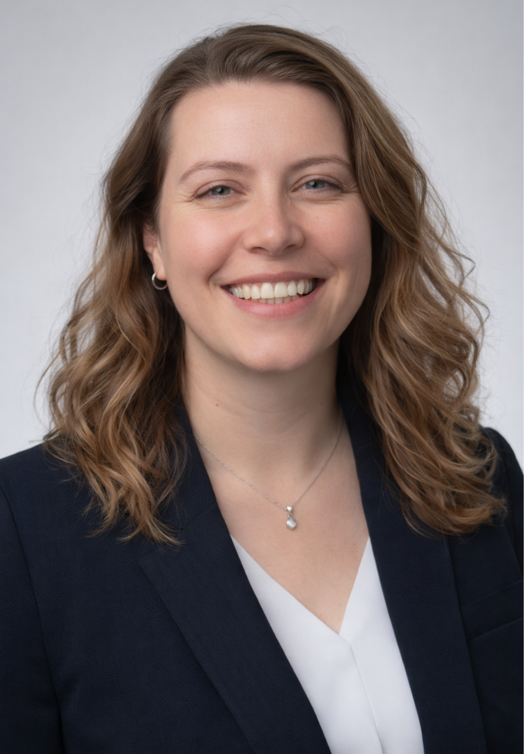 A smiling white woman with wavy, shoulder-length brown hair, wearing a black blazer, white blouse, silver necklace, and hoop earrings, against a light gray background.