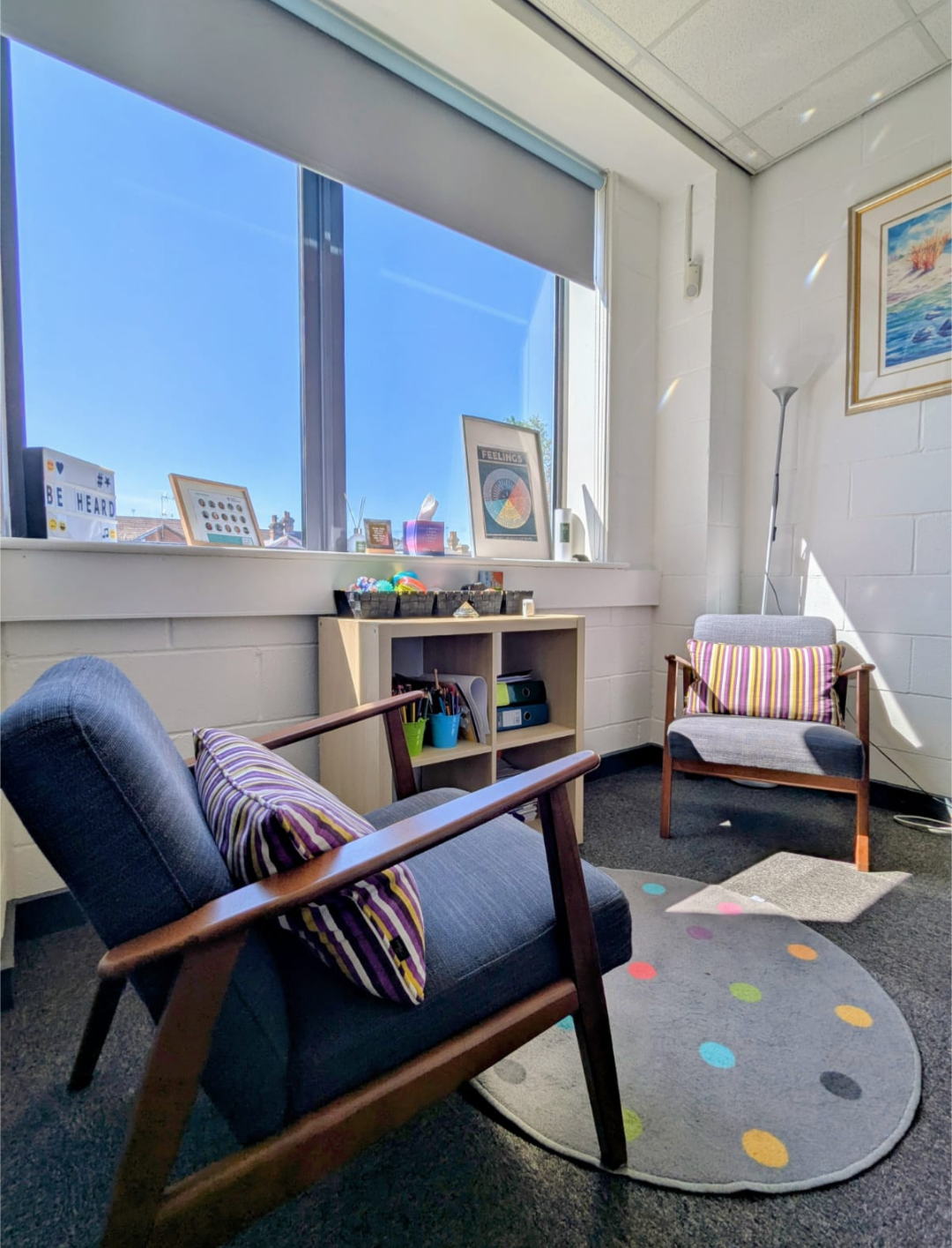 A cozy, sunlit office with two chairs, a small bookshelf, colorful cushions, a circular rug with colorful dots, a large window, artwork on the wall, and various decorations on the windowsill.