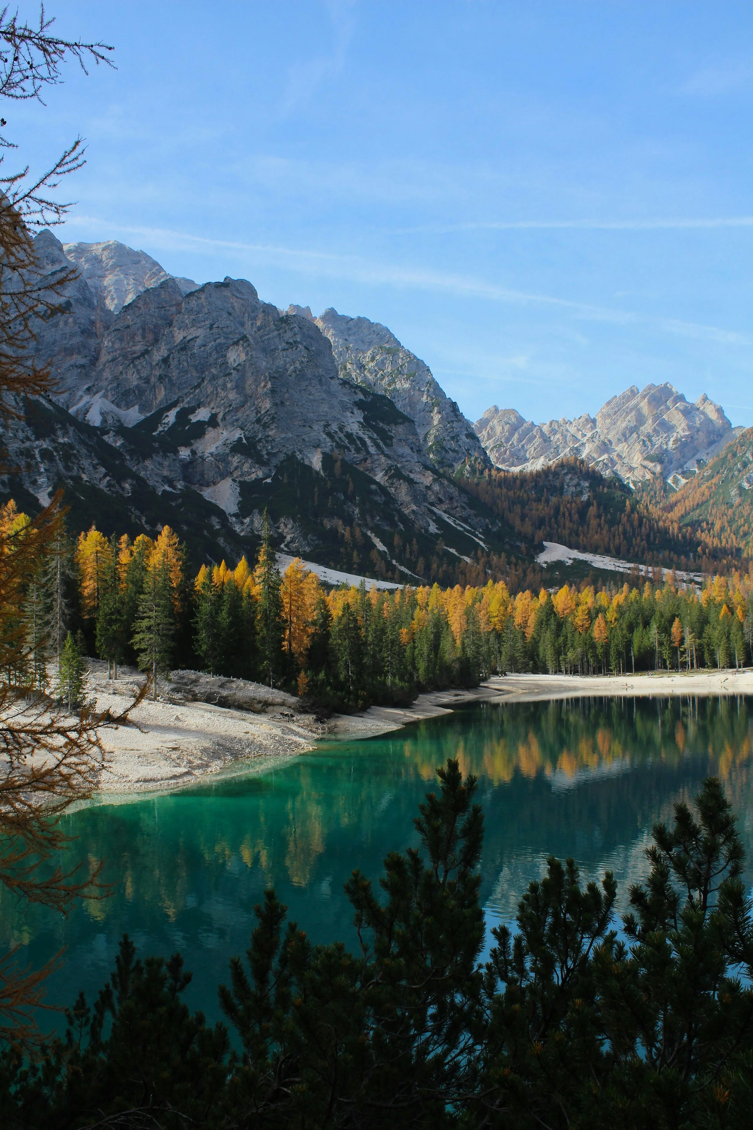 A serene mountain landscape with rocky peaks, a forest of pine trees, and a calm turquoise lake reflecting the scenery, under a clear blue sky.