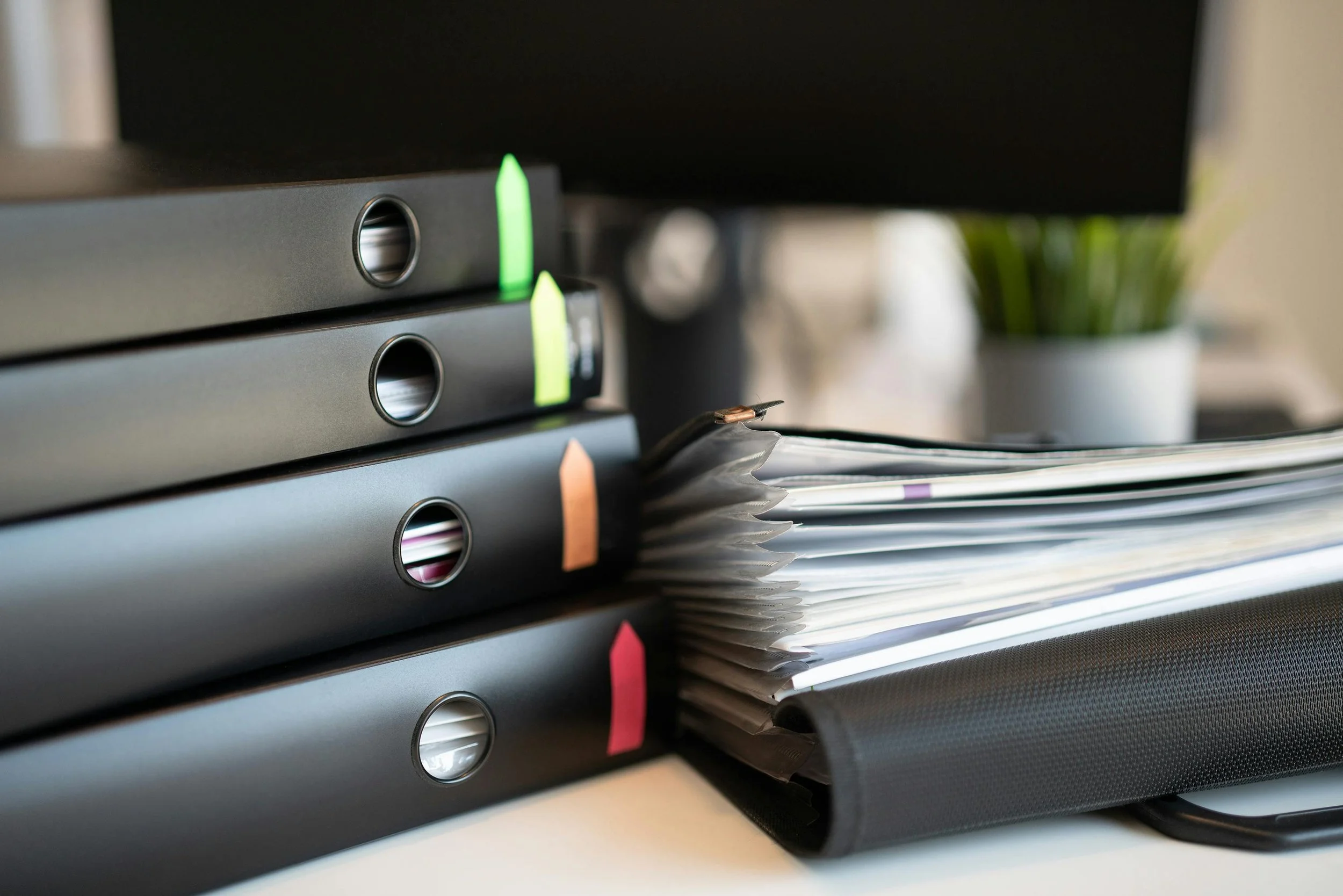Stacked black binders with colorful sticky notes on a desk, with papers and a plant in the background.