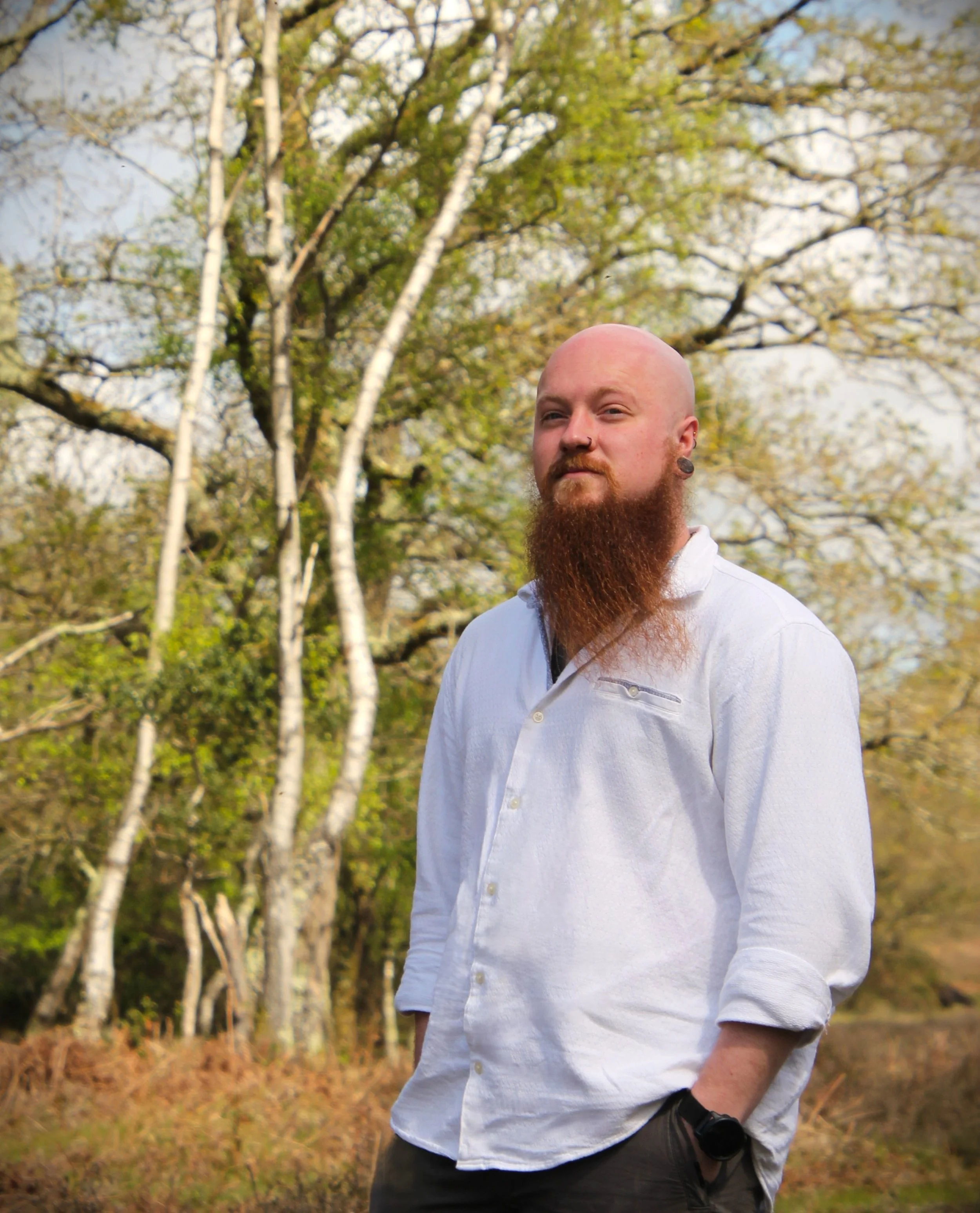 Bald man with a beard, earrings, and a nose piercing, wearing a white shirt, standing outdoors near trees with budding leaves in spring.
